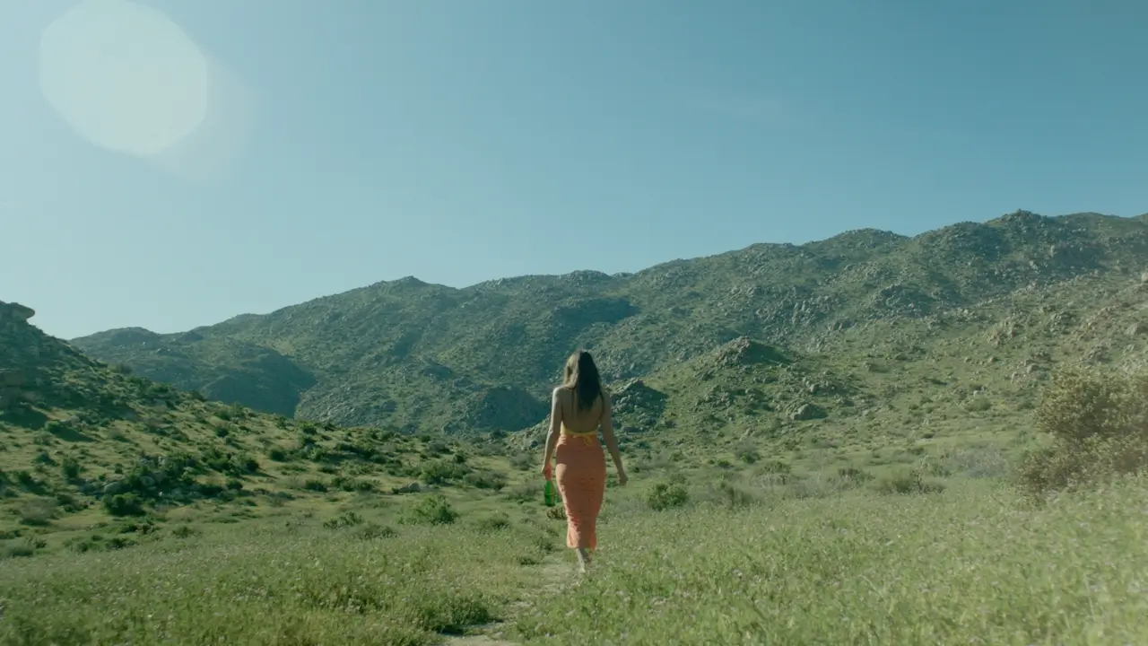 Woman in an orange skirt walking barefoot on a grassy path toward green rocky mountains under a clear blue sky.