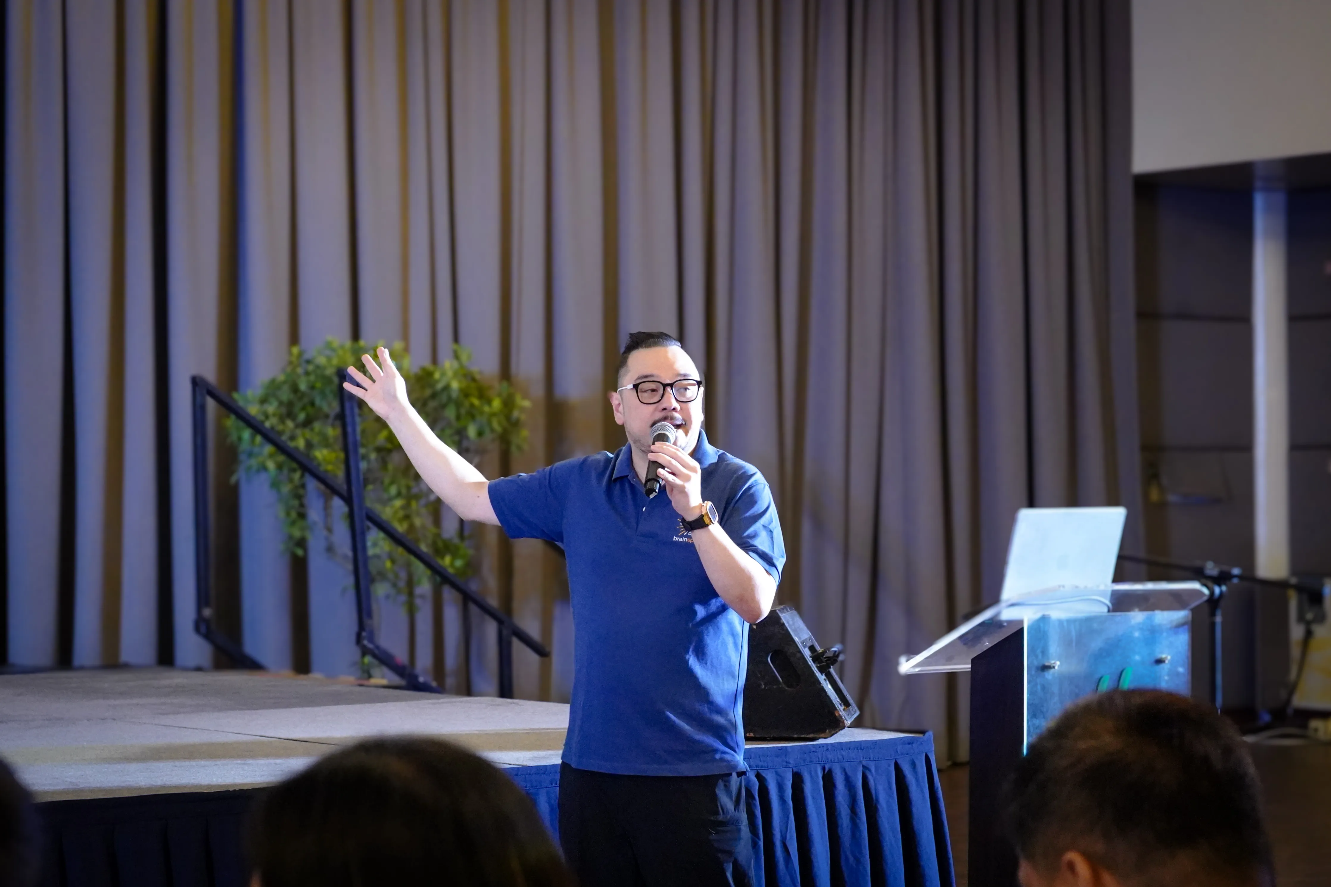 Man wearing glasses and a blue polo shirt speaking into a microphone on a stage with a laptop and podium nearby.