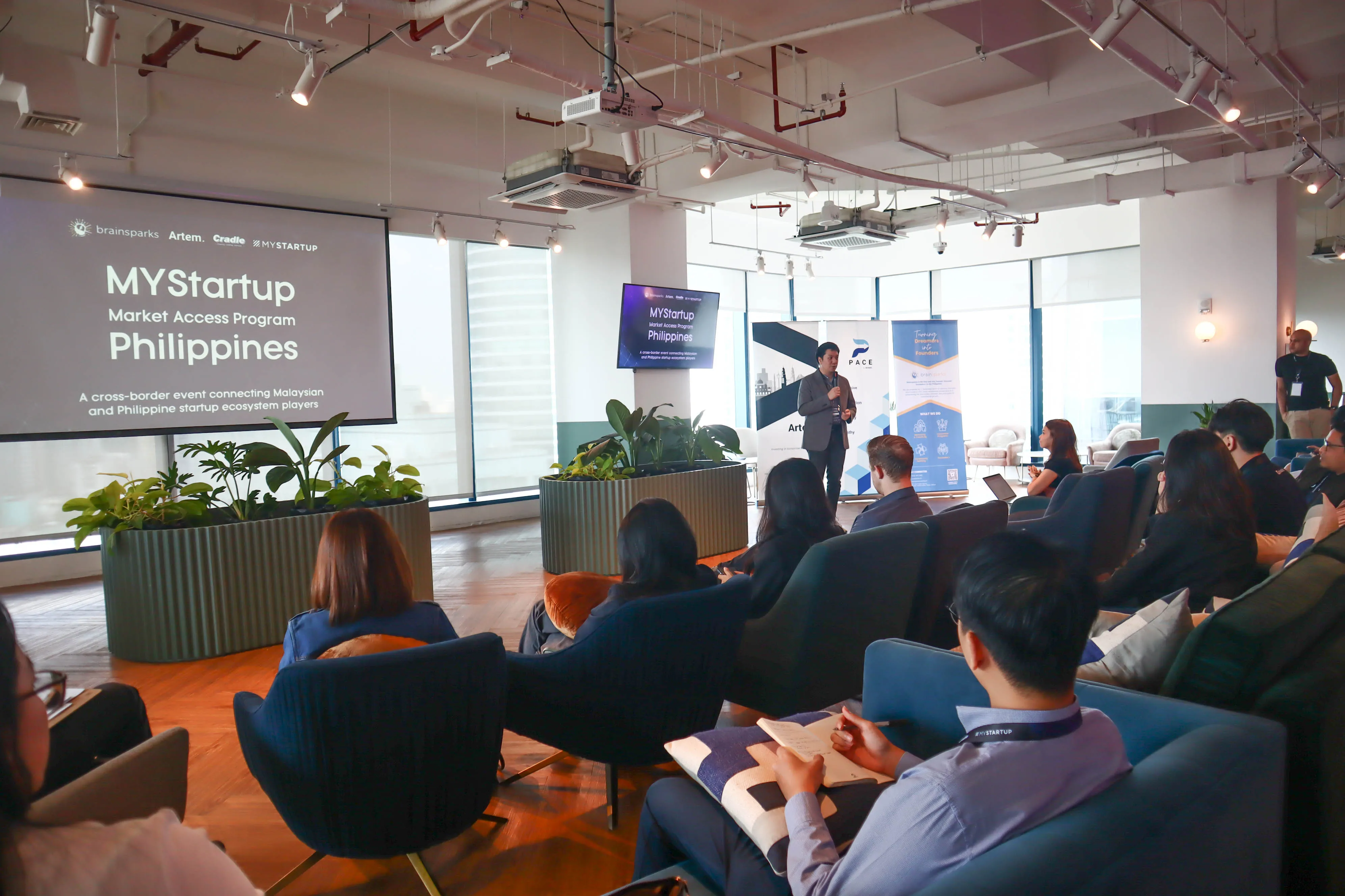 Man in a blazer giving a presentation to an audience seated in a modern conference room with a screen displaying MYStartup Market Access Program Philippines.