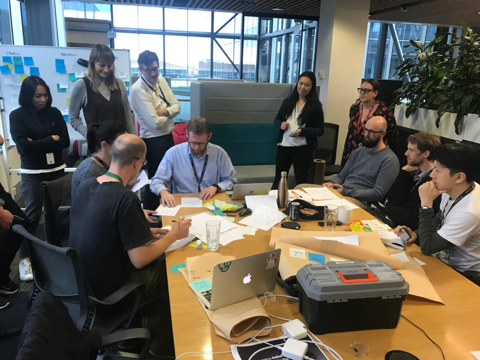 A group of people engaged in a meeting around a table with laptops, papers, and a toolbox in a modern office.