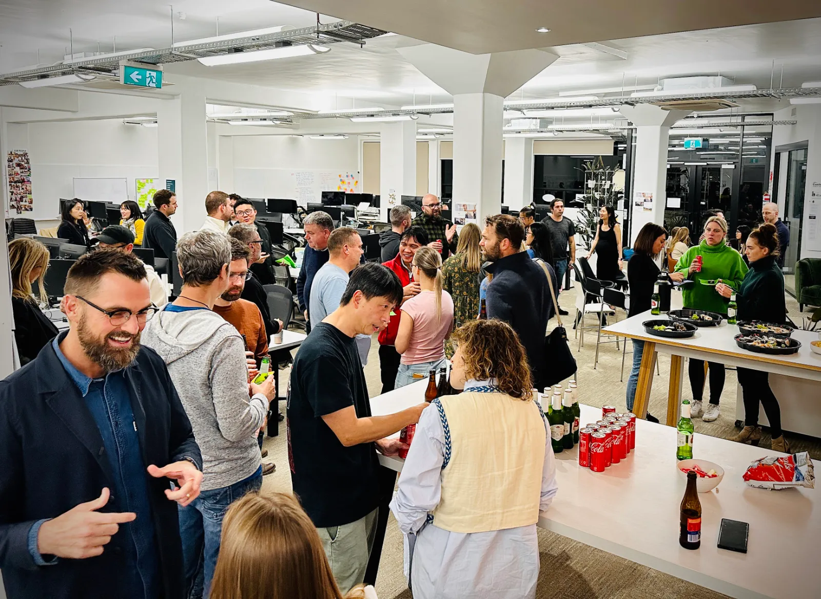 Office gathering with people socializing, holding drinks, and snacks on tables in a bright open workspace.