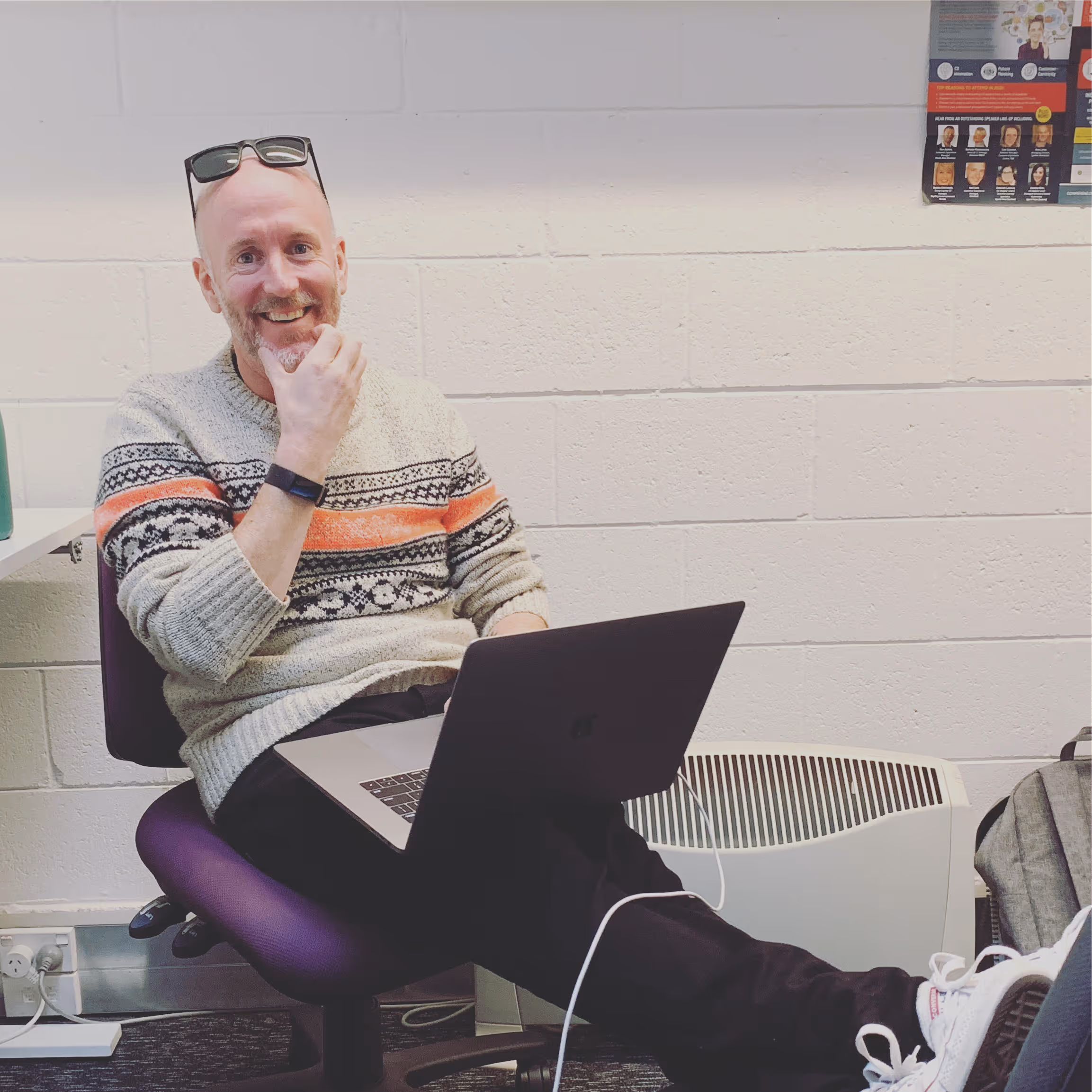 Smiling man wearing sunglasses on head and patterned sweater, sitting on a purple chair with a laptop on his lap and feet resting on a table.