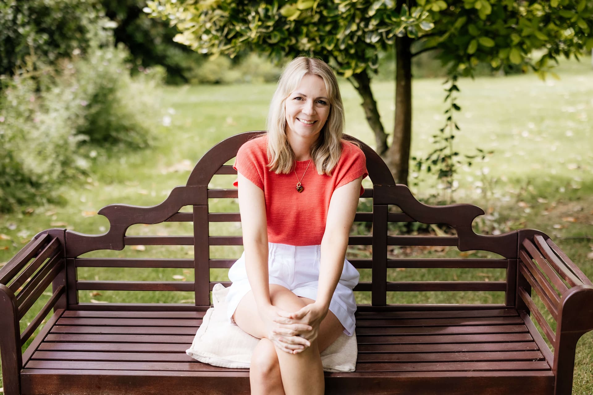 Smiling blonde woman wearing a red top and white skirt sitting on a wooden bench outdoors with greenery in the background.