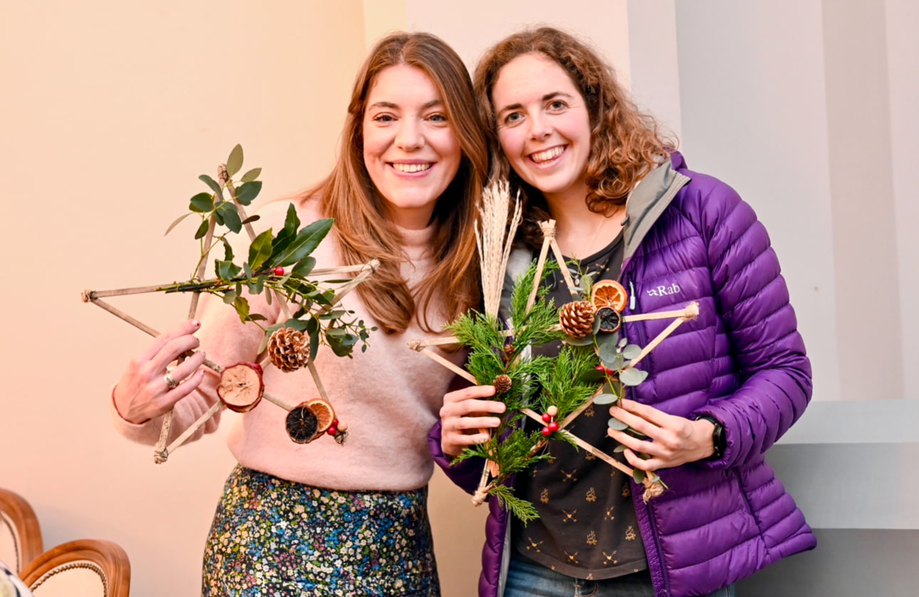 Two smiling women holding star-shaped decorations made of twigs, pinecones, dried citrus, and greenery.