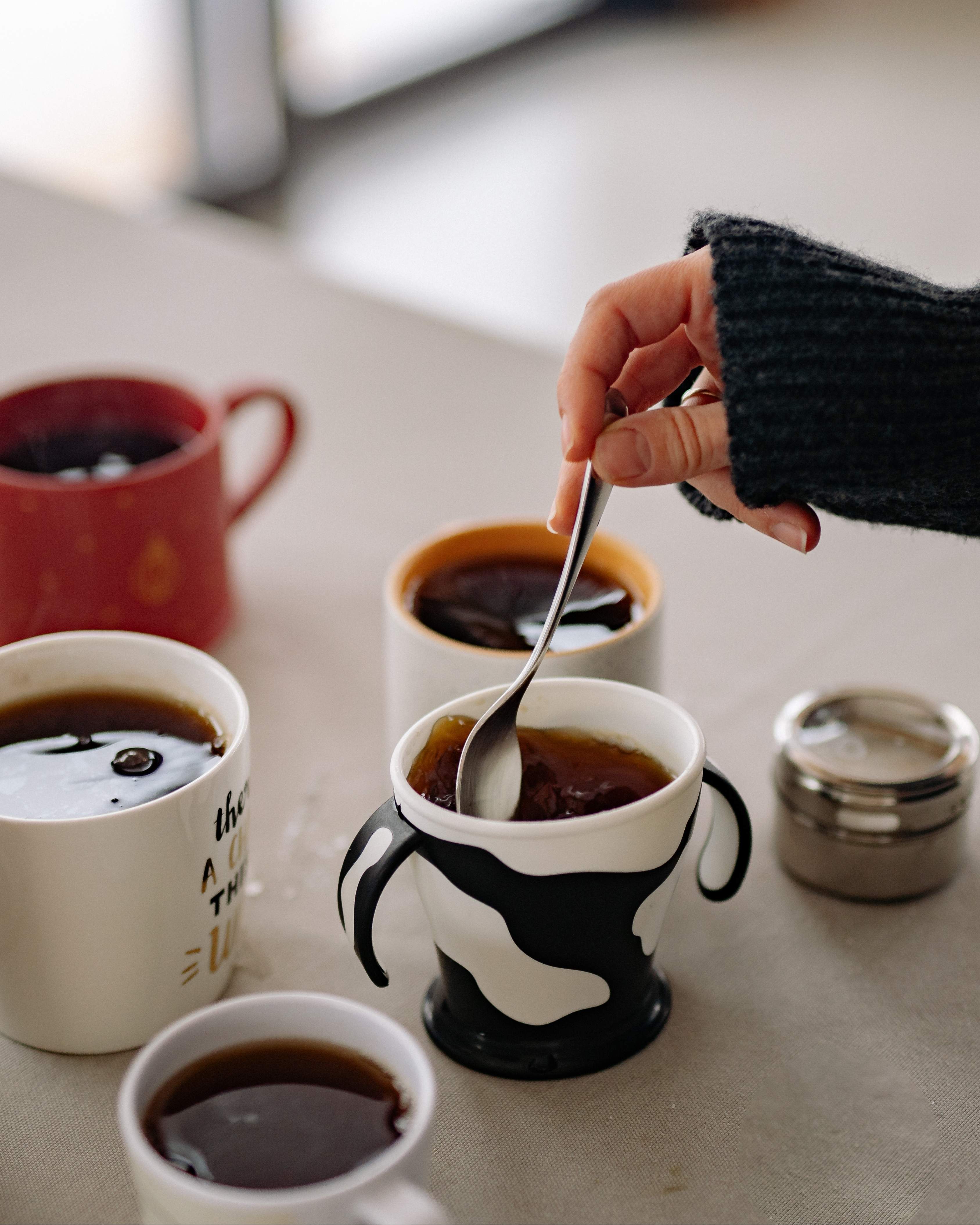 Hand stirring coffee in a black and white cow-patterned mug surrounded by four other coffee mugs on a table.