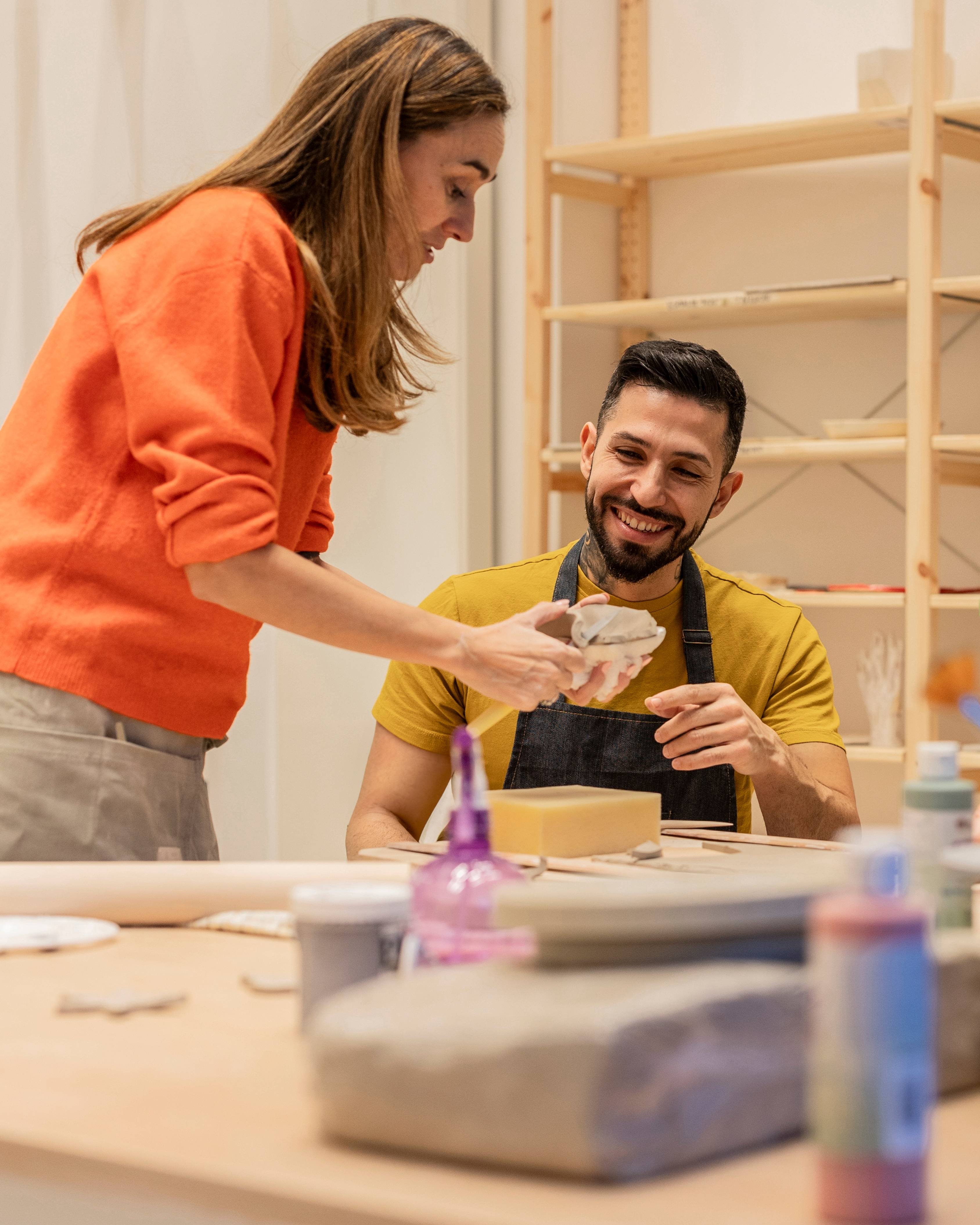 Woman in orange sweater showing a piece of clay to a smiling man wearing a yellow shirt and black apron in a pottery studio.