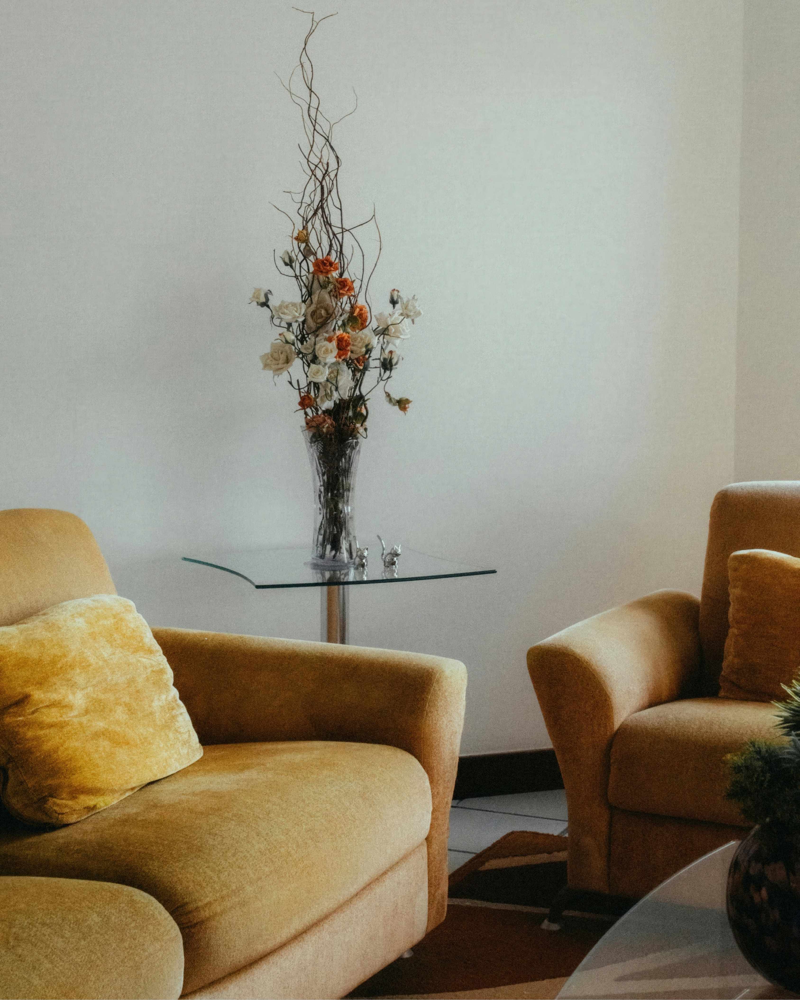 Two beige armchairs with yellow cushions flank a glass table holding a vase of dried flowers.