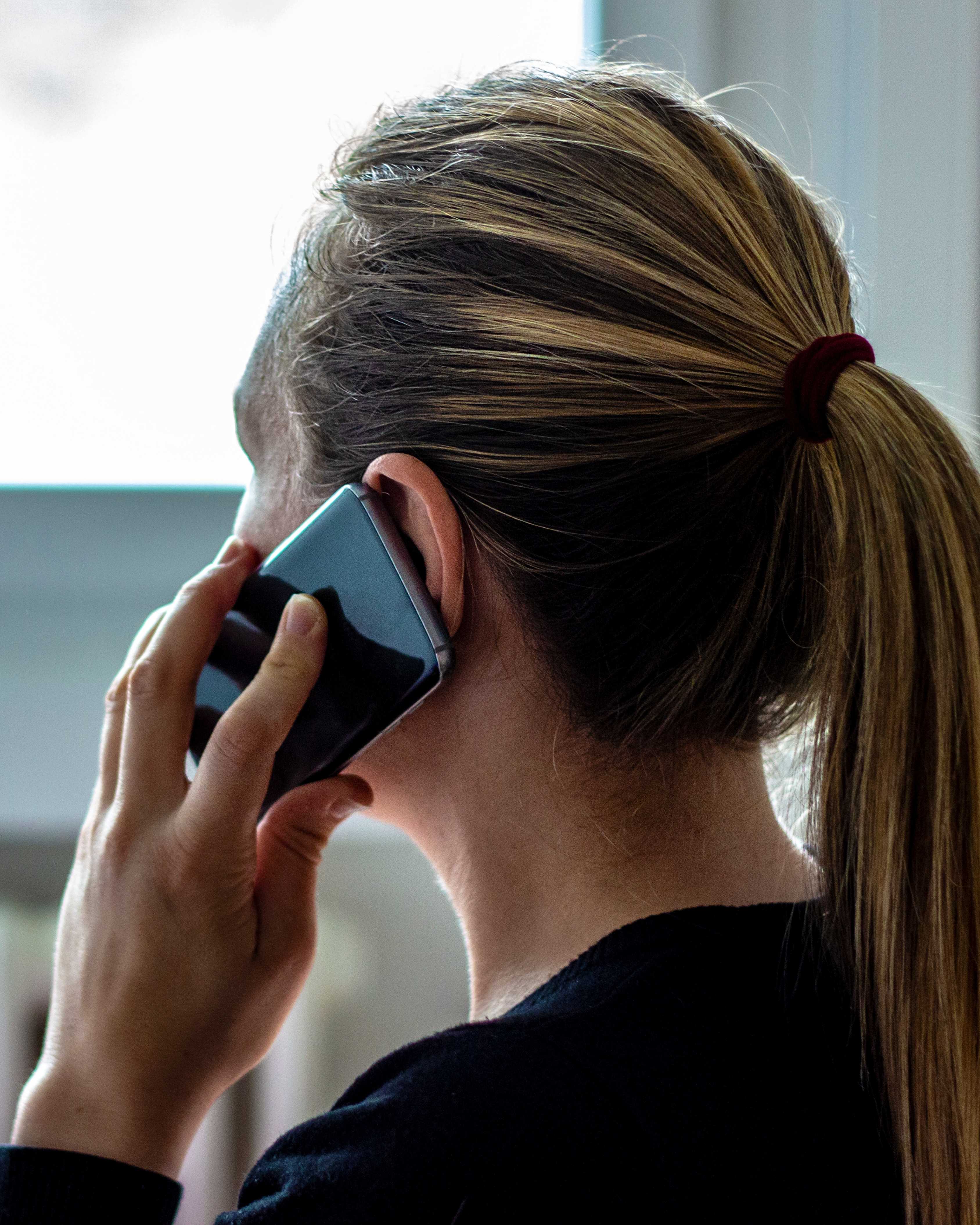 Person with blonde hair tied in a ponytail holding a smartphone to their ear while talking.
