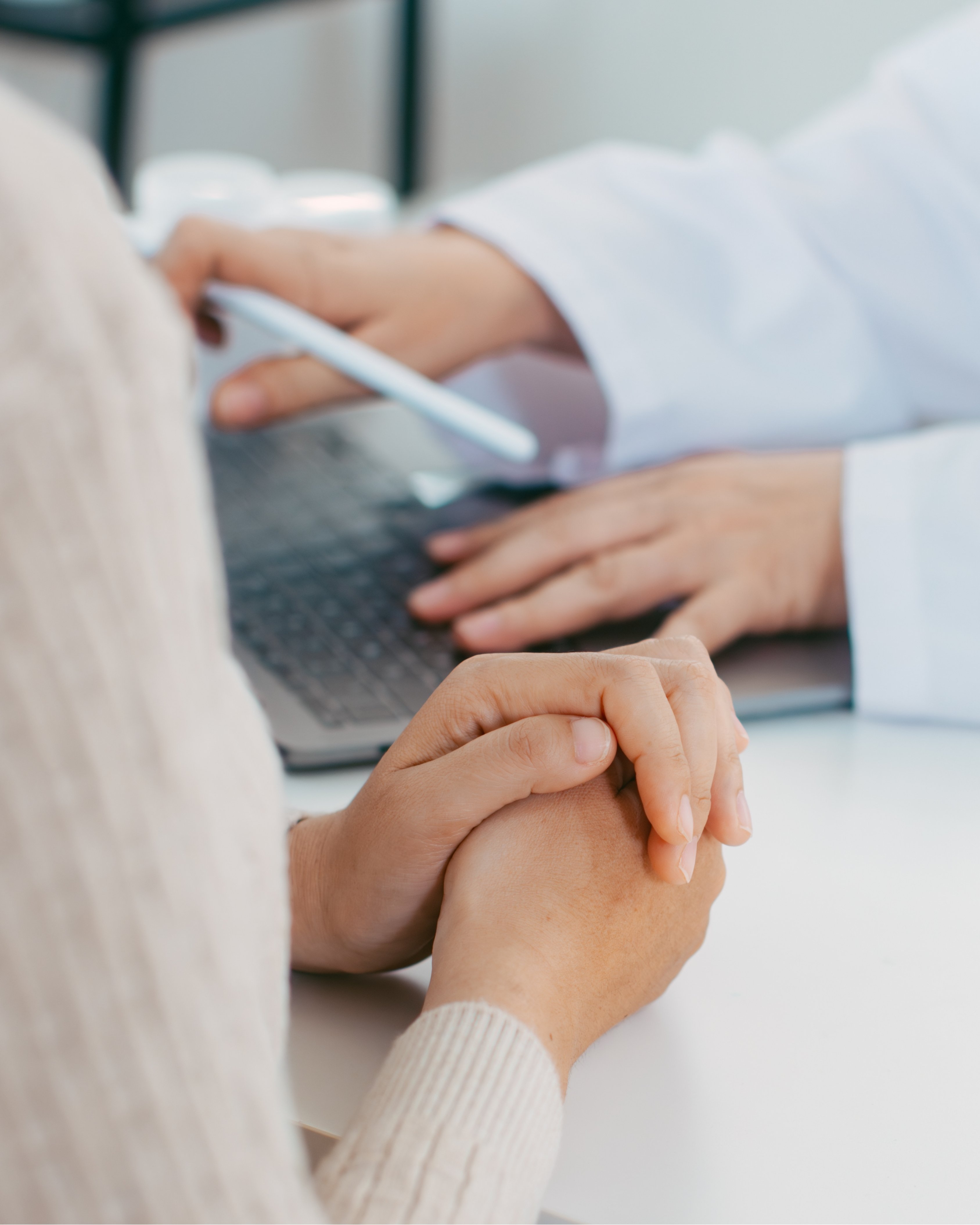 Close-up of a person holding their own hands on a desk while a healthcare professional uses a stylus on a laptop keyboard.
