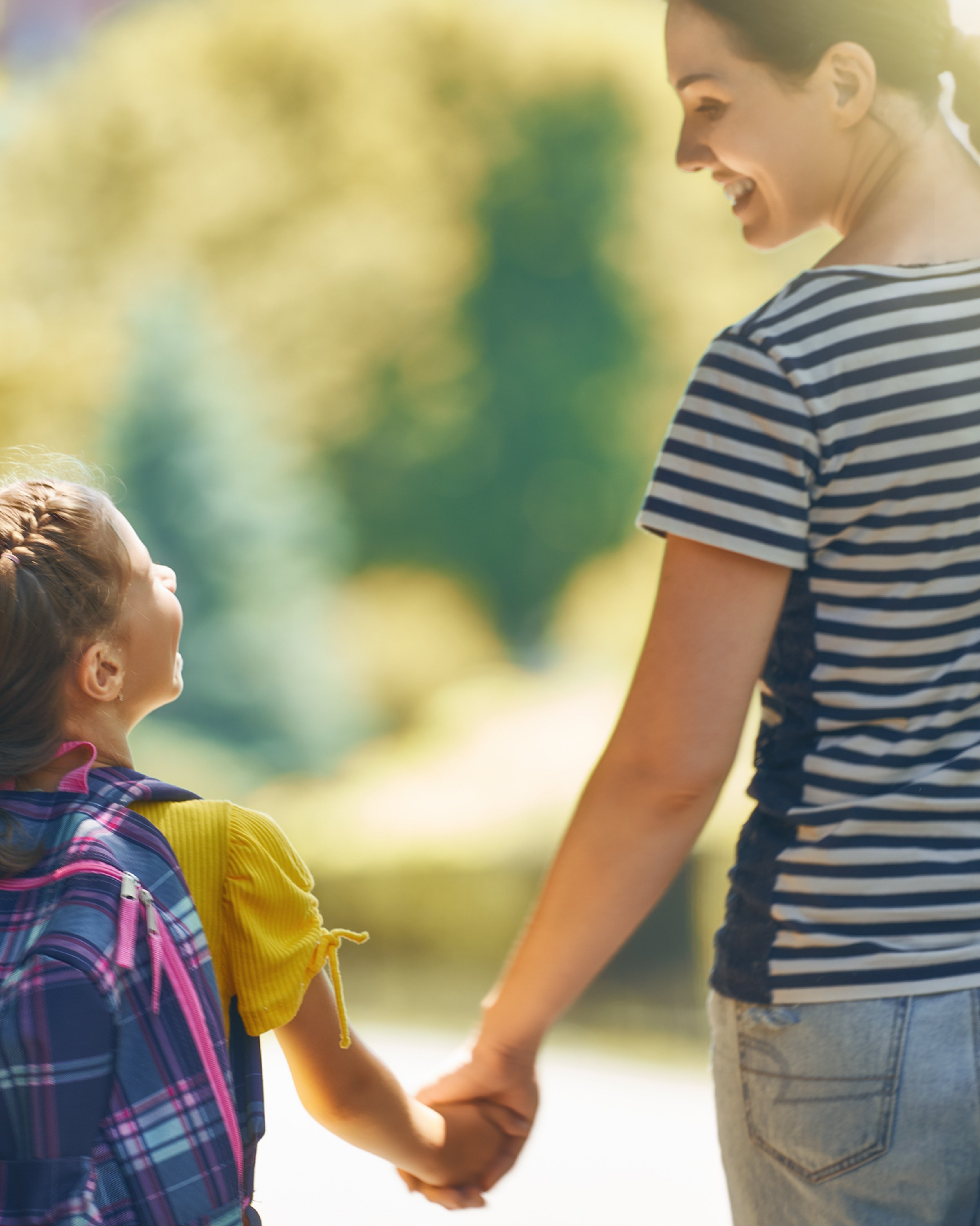 Smiling woman holding hands with a young girl carrying a backpack, walking outdoors on a sunny day.