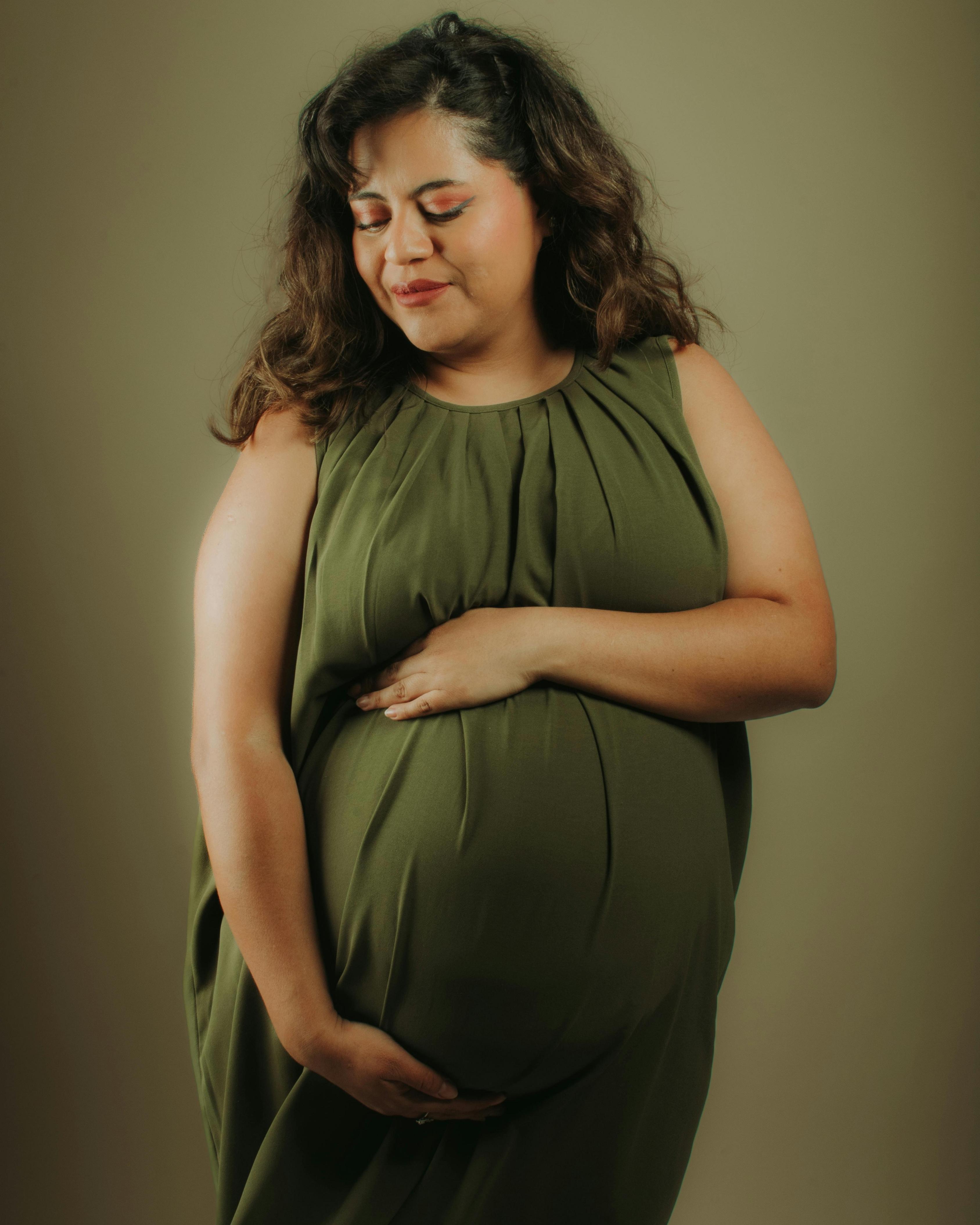 Pregnant woman in a green dress gently cradling her belly and looking down with a soft smile.