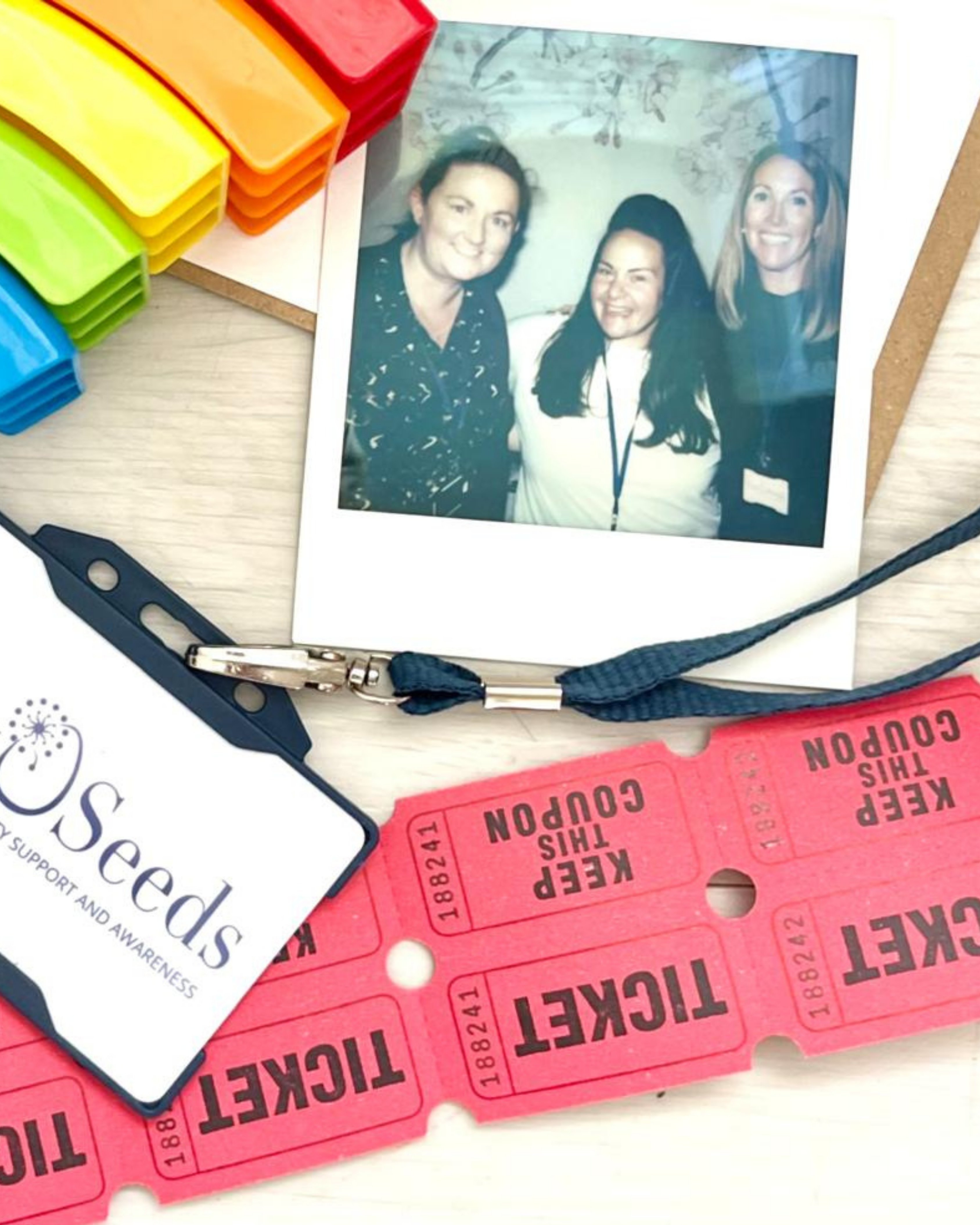 Polaroid photo of three smiling women surrounded by colorful plastic clips, a volunteer badge labeled Seeds, and red raffle tickets.