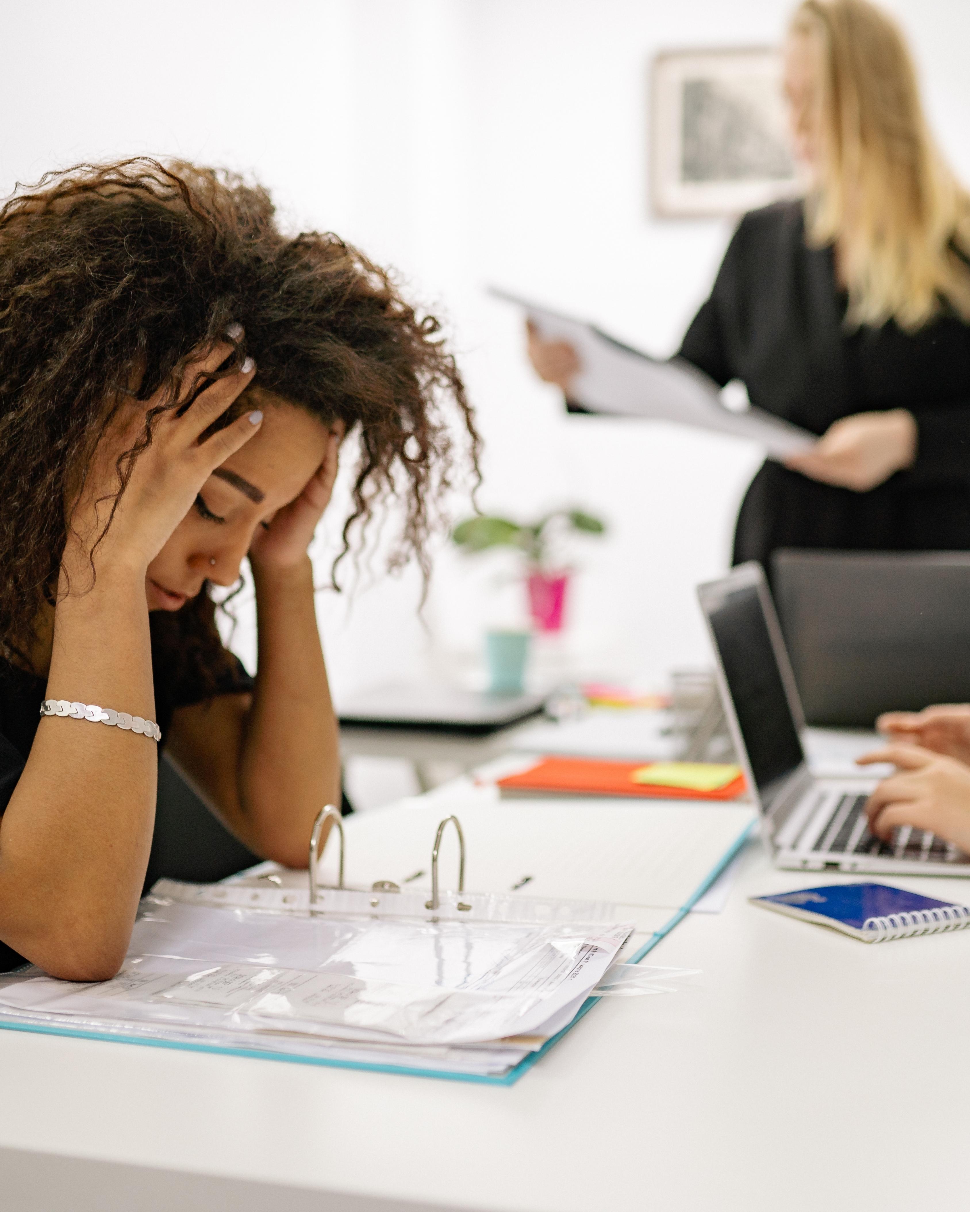 Stressed woman sitting at a desk with head in hands and open binder, while a colleague works on a laptop in a bright office.