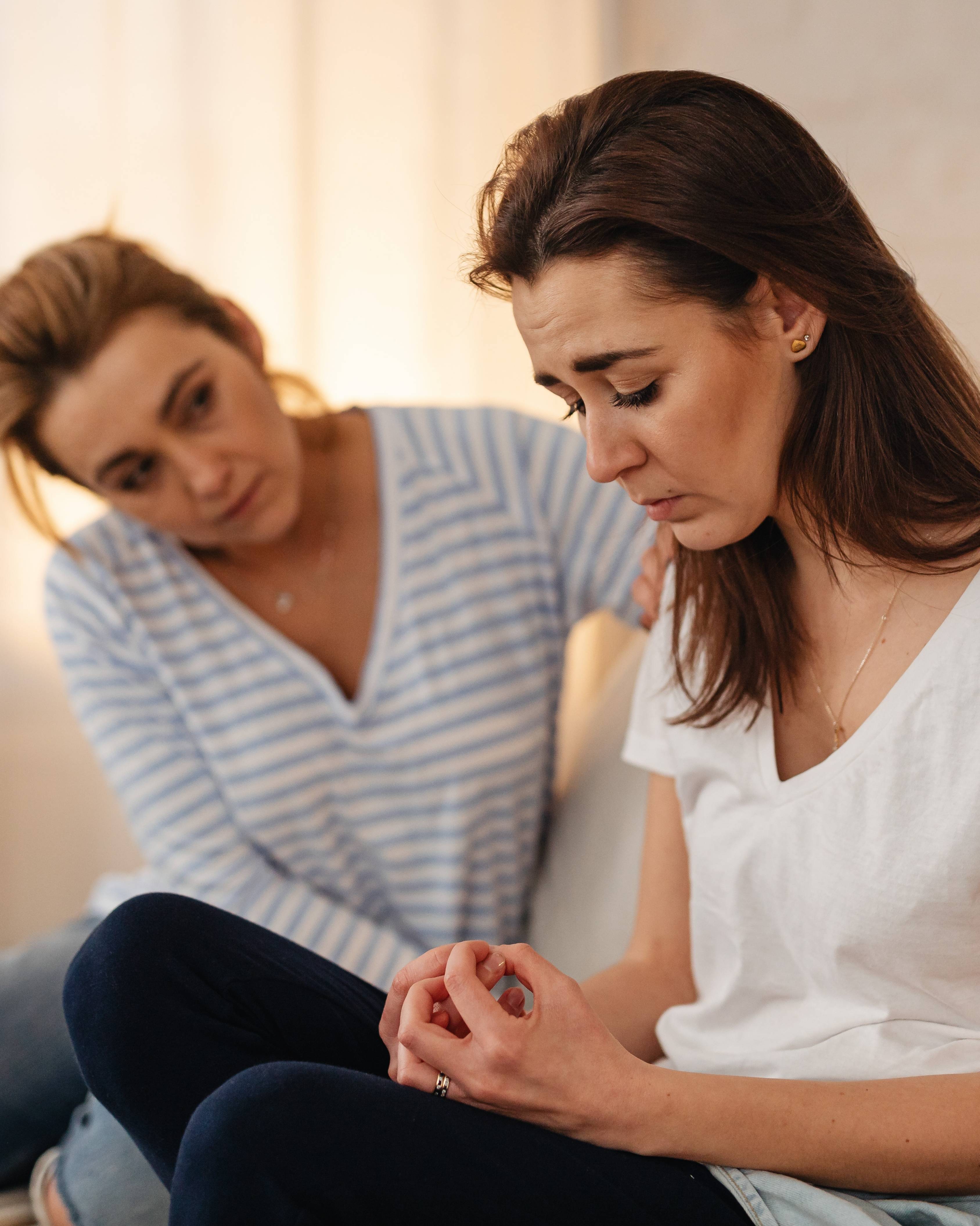 Woman in white t-shirt looking distressed while another woman in striped shirt comforts her by placing a hand on her shoulder.