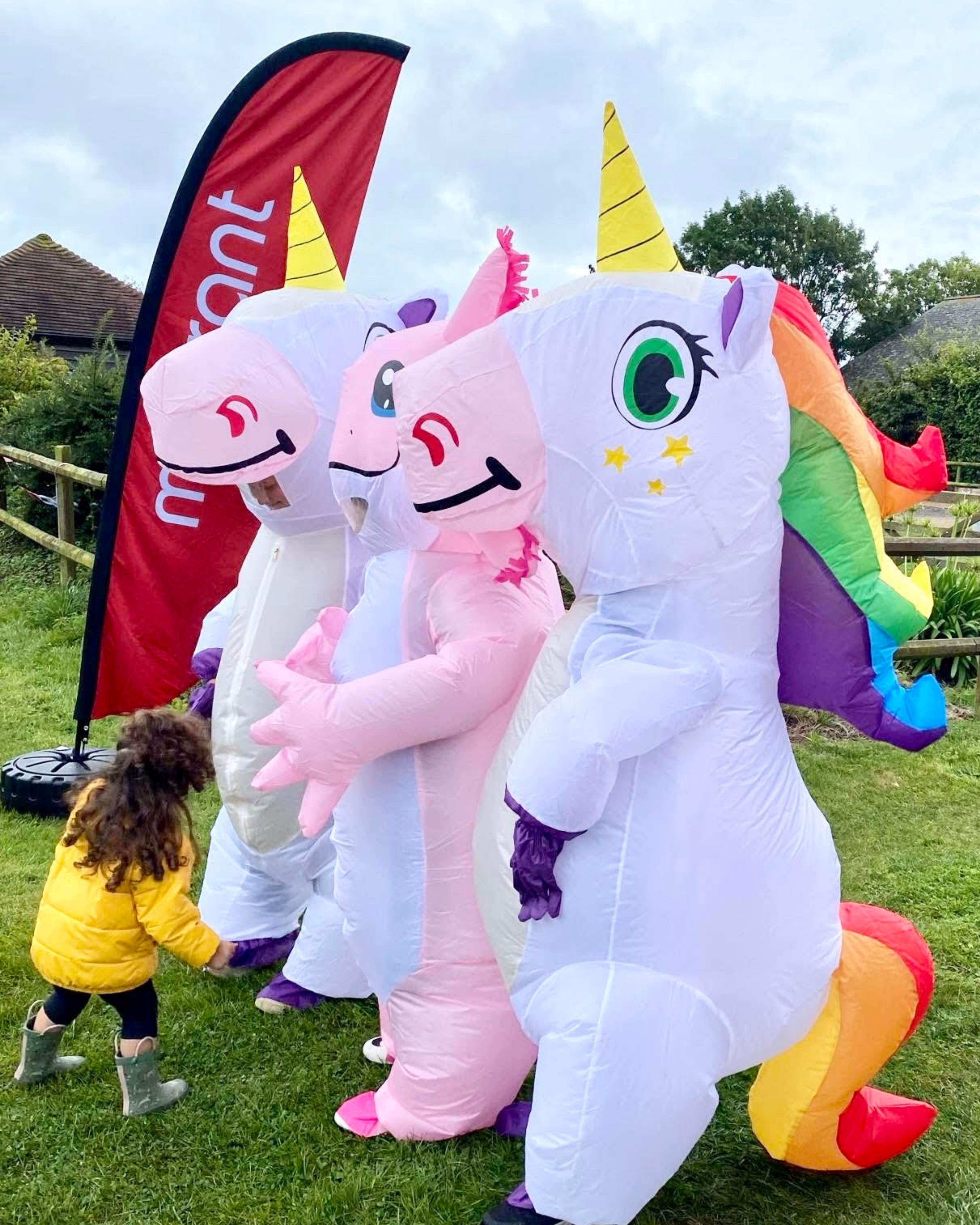 Child in yellow coat and boots reaching out to three people in white and pink inflatable unicorn costumes on green grass.