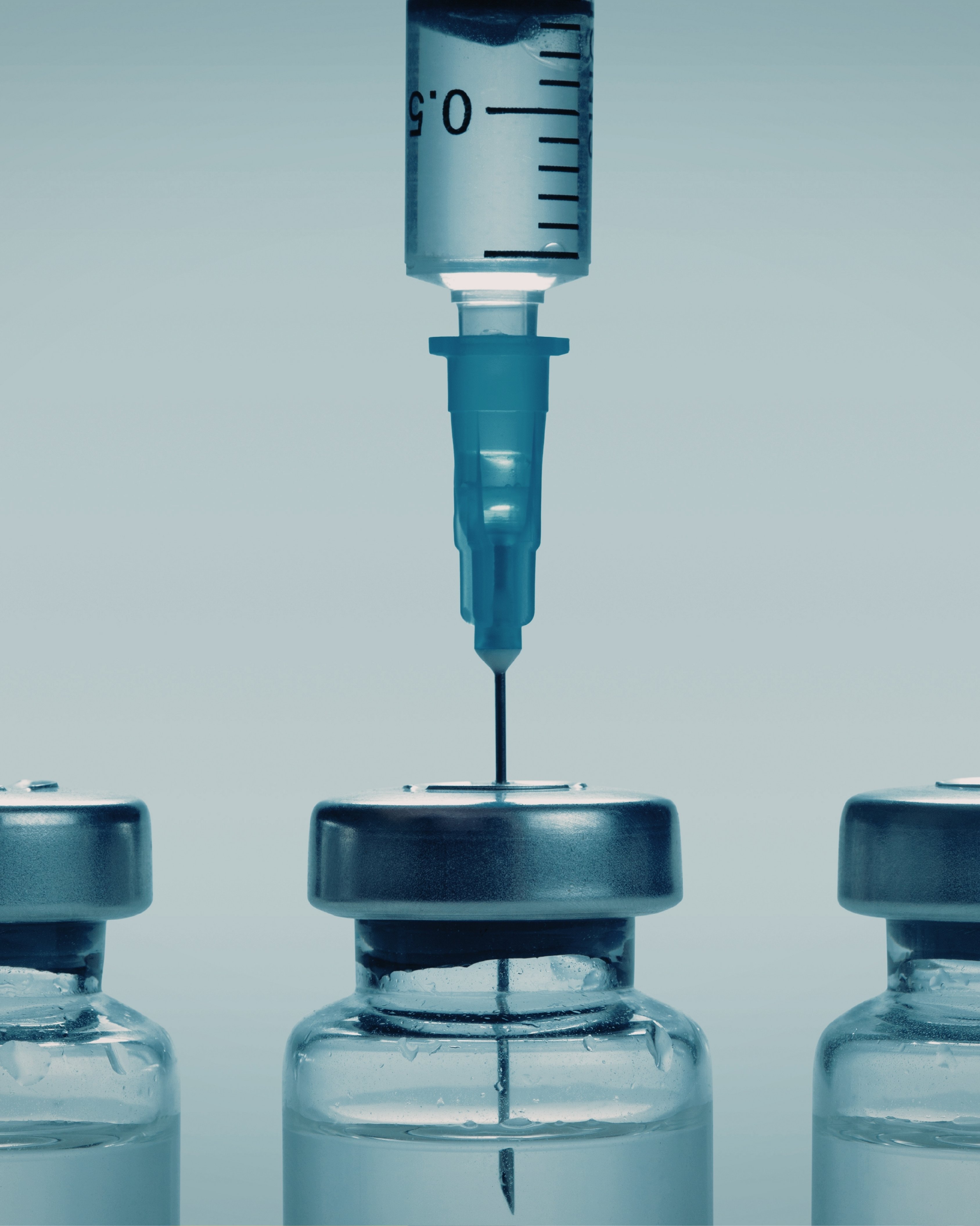 Syringe needle inserted into the rubber stopper of a vaccine vial, with two additional vials partially visible on either side.