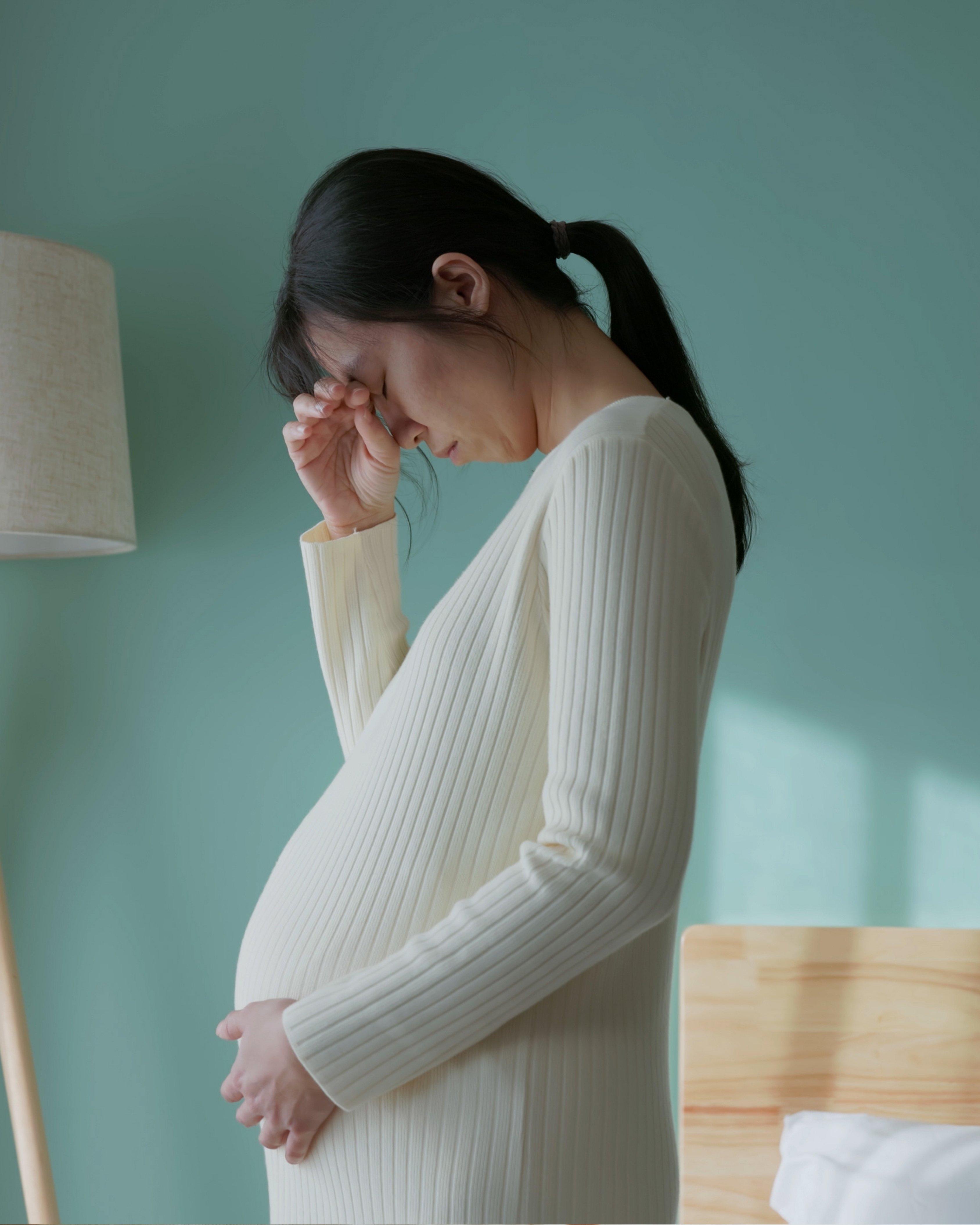 Pregnant woman in a white ribbed dress holding her belly and touching her forehead with a concerned expression.
