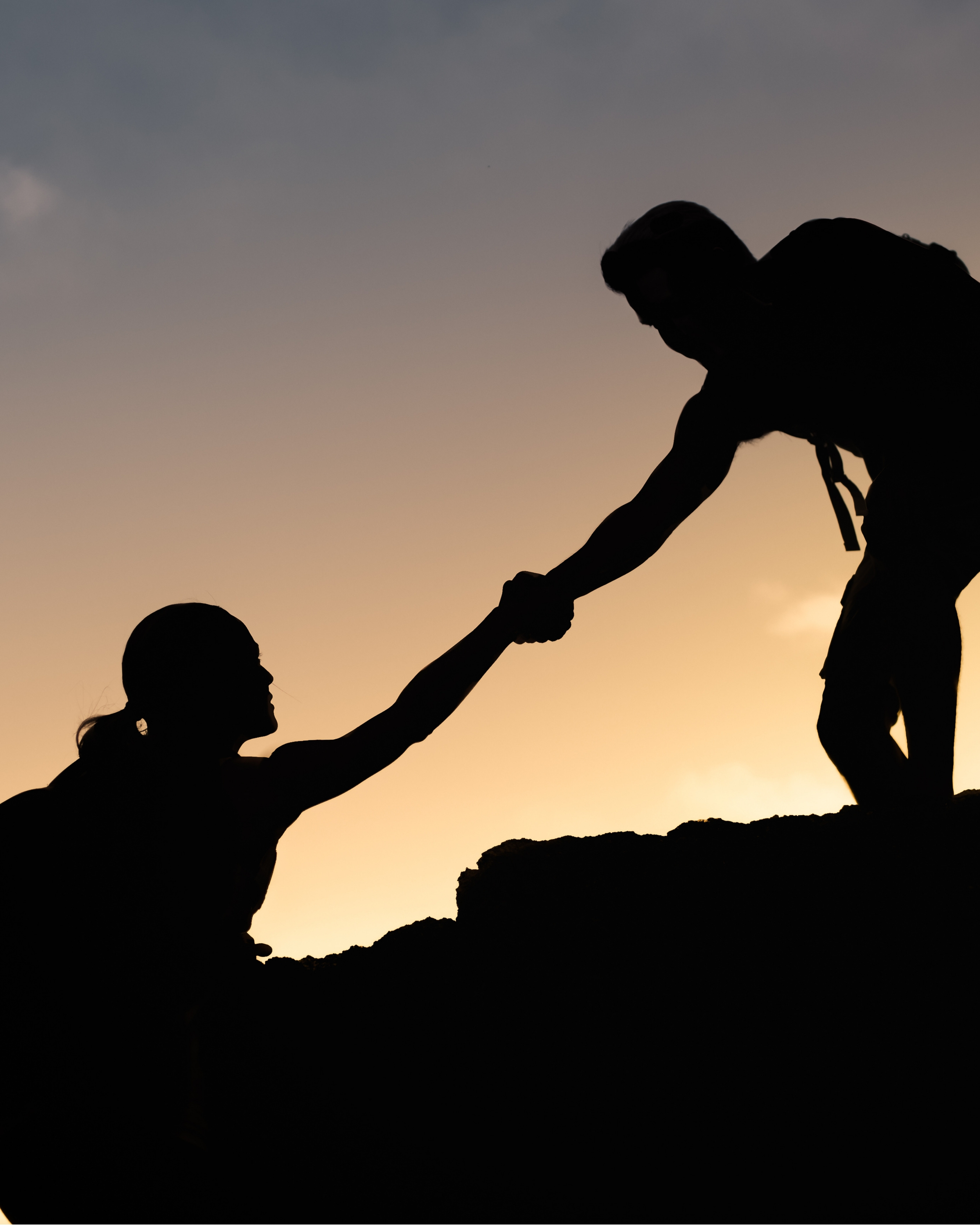 Silhouette of a person helping another climb up a rocky slope at sunset.