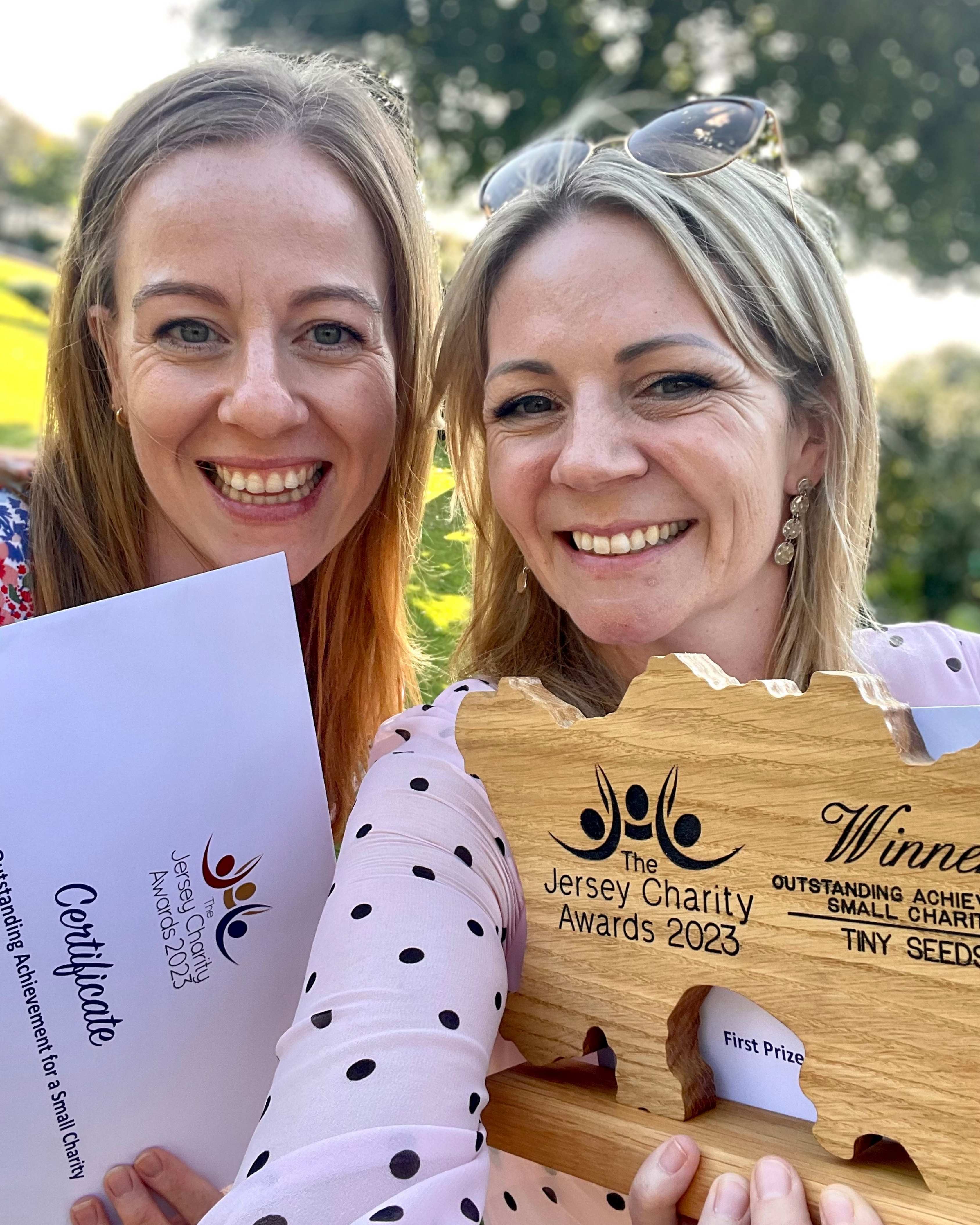 Two smiling women holding a wooden award and certificate for Outstanding Achievement for Small Charity at The Jersey Charity Awards 2023.