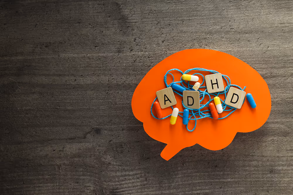 En orange hjärnformad platta ligger på ett bord med bokstavsklossar som bildar ordet ADHD och flera kapslar, som illustrerar medicinsk behandling och individuell uppföljning hos Vigilans Umeå AB.