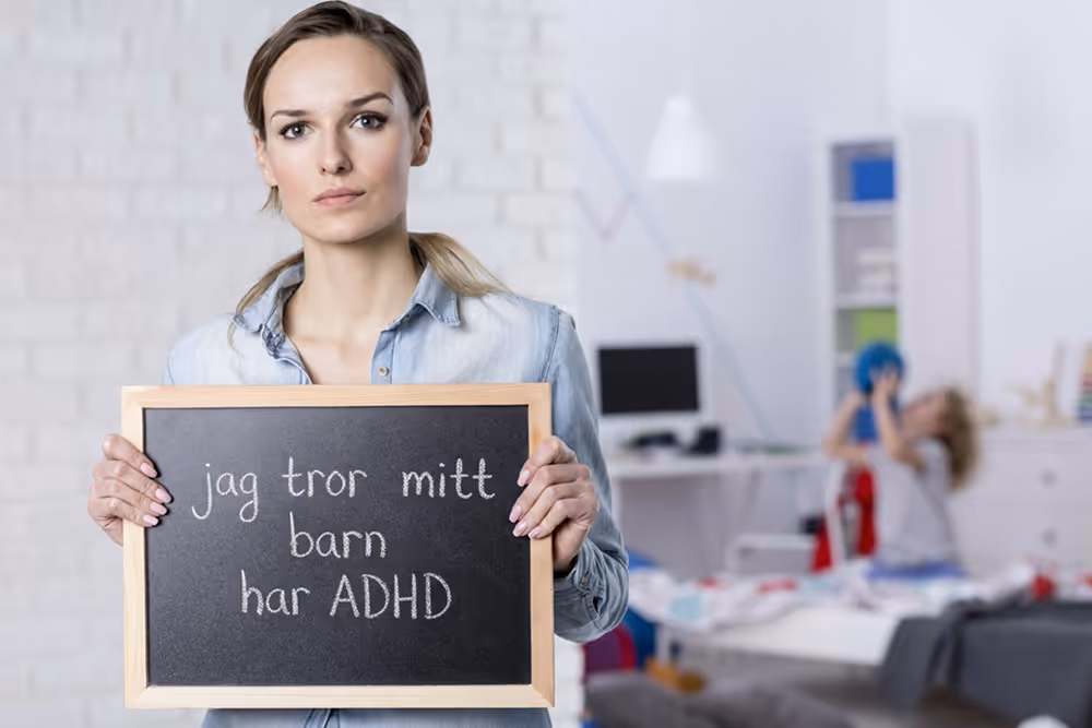 En förälder håller en griffeltavla med texten ”jag tror mitt barn har ADHD” i ett barnrum, vilket speglar behovet av tydlig och snabb ADHD-utredning hos Vigilans Umeå AB.