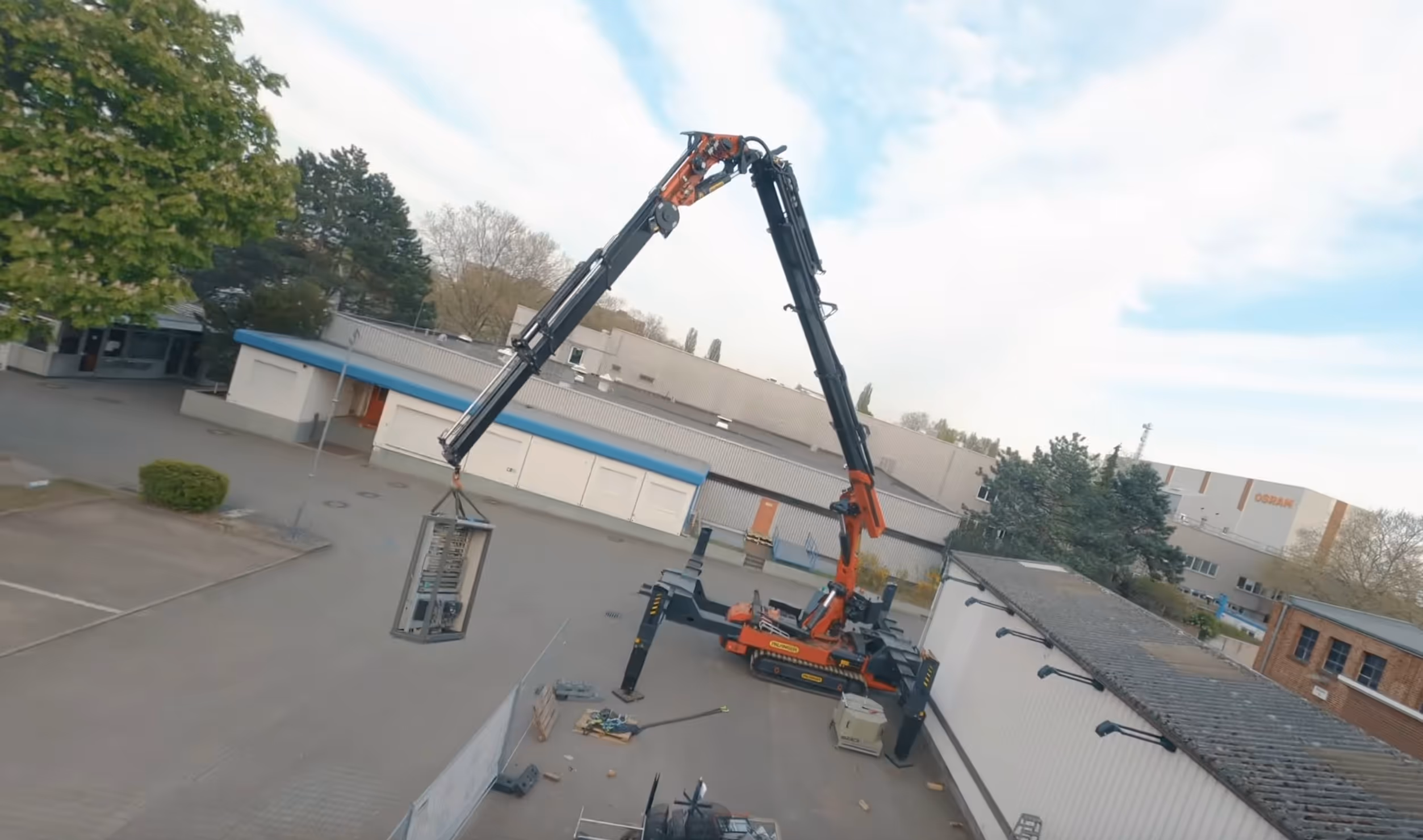 Large red and black crane with extended arm lifting a metal crate in an industrial outdoor area.