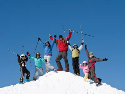 Groupe de six personnes, dont des enfants, debout et célébrant au sommet d'une colline de neige avec un ciel bleu clair en arrière-plan.