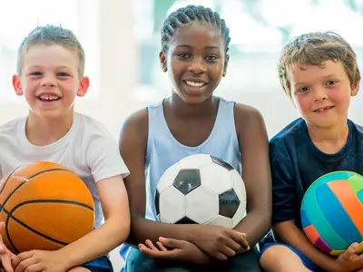Trois enfants souriants, chacun tenant un ballon différent : un ballon de basket, un ballon de football et un ballon coloré.