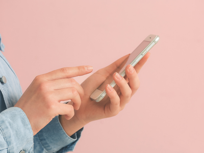 Une personne tenant un smartphone argenté avec une main et utilisant l'autre pour toucher l'écran, sur fond rose.