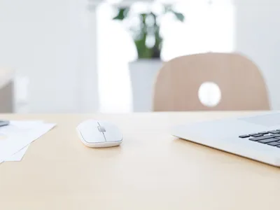 Souris blanche et partie d'un ordinateur portable sur un bureau en bois clair avec une chaise floue et une plante en arrière-plan.