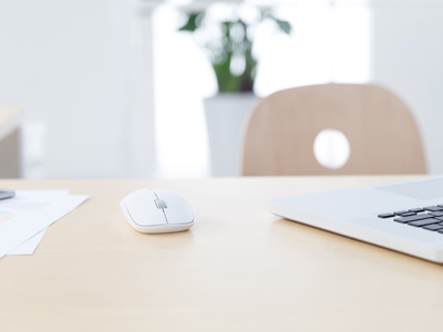 Souris blanche et partie d'un ordinateur portable sur un bureau en bois clair avec une chaise floue et une plante en arrière-plan.