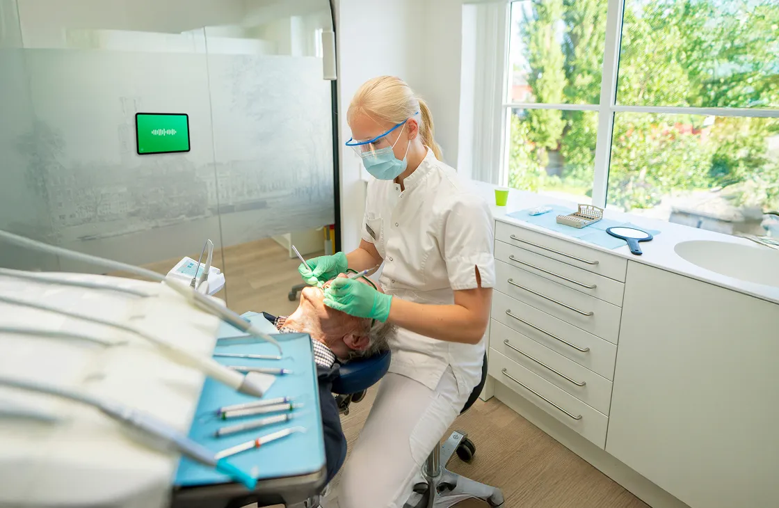 Dentist wearing protective eyewear and mask performing dental checkup on patient in a bright clinic room with large window showing greenery outside.