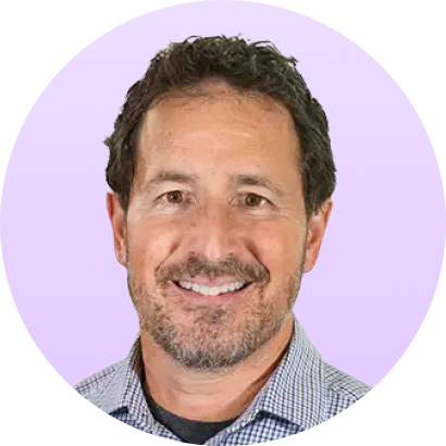 Smiling middle-aged man with short curly dark hair and a trimmed beard wearing a checkered shirt against a light purple background.