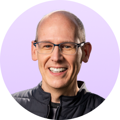 Smiling bald man with glasses wearing a black jacket against a light purple background.