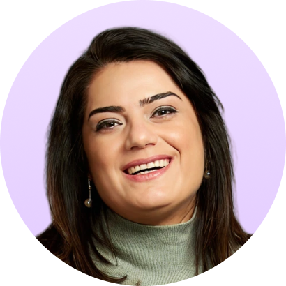 Smiling woman with dark hair wearing a green turtleneck sweater and pearl earrings on a light purple background.