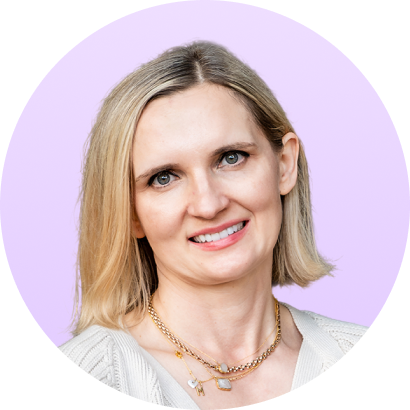 Smiling blonde woman with short hair wearing layered gold necklaces and a white sweater against a light purple background.