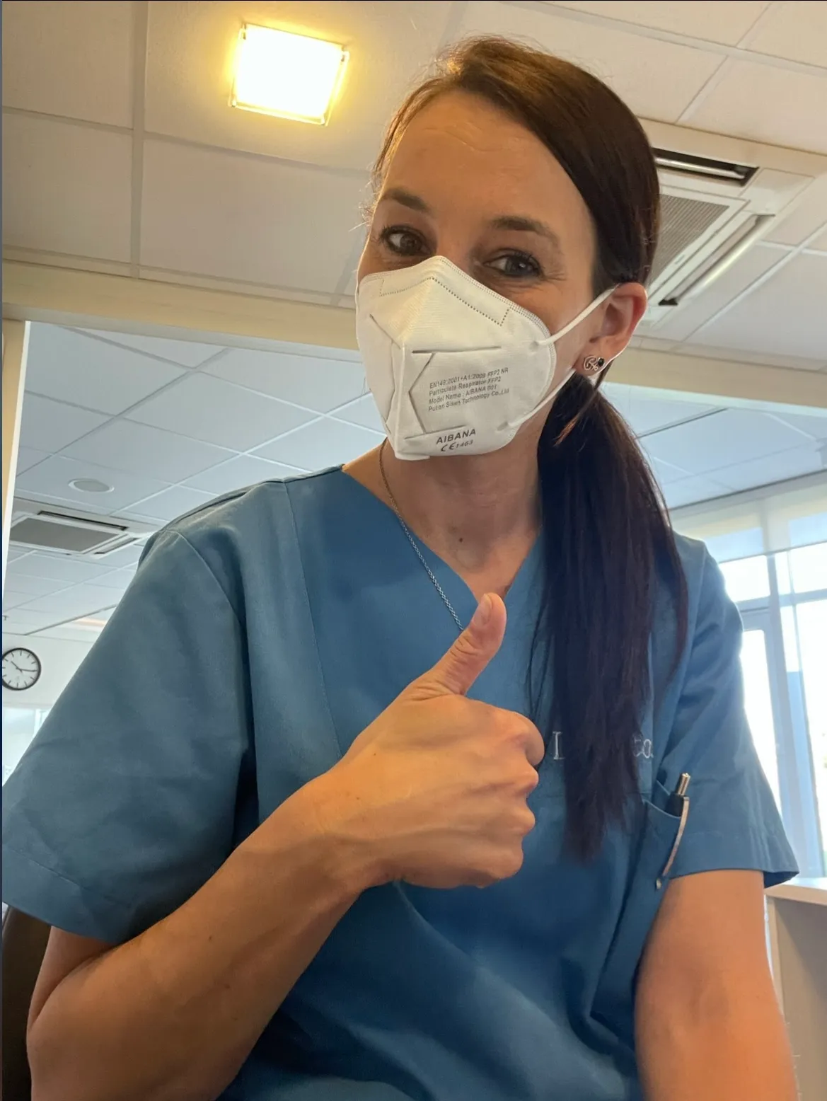 Frau in medizinischer Schutzmaske und blauer Arbeitskleidung zeigt Daumen hoch in einem hellen Innenraum mit Decke und Fenstern.