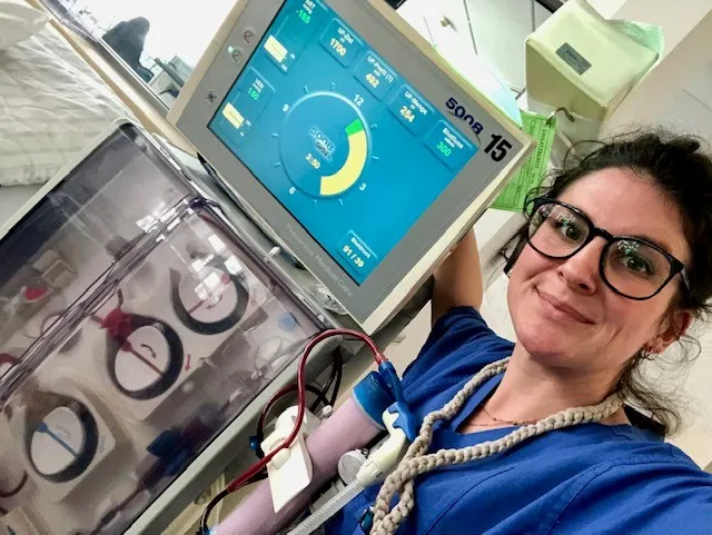 Eine Frau in blauer medizinischer Kleidung und Brille macht ein Selfie neben einem medizinischen Gerät mit einem Bildschirm, der Vitaldaten und einen Kreisdiagramm anzeigt; sie hat einen Schlauch am Arm verbunden.
