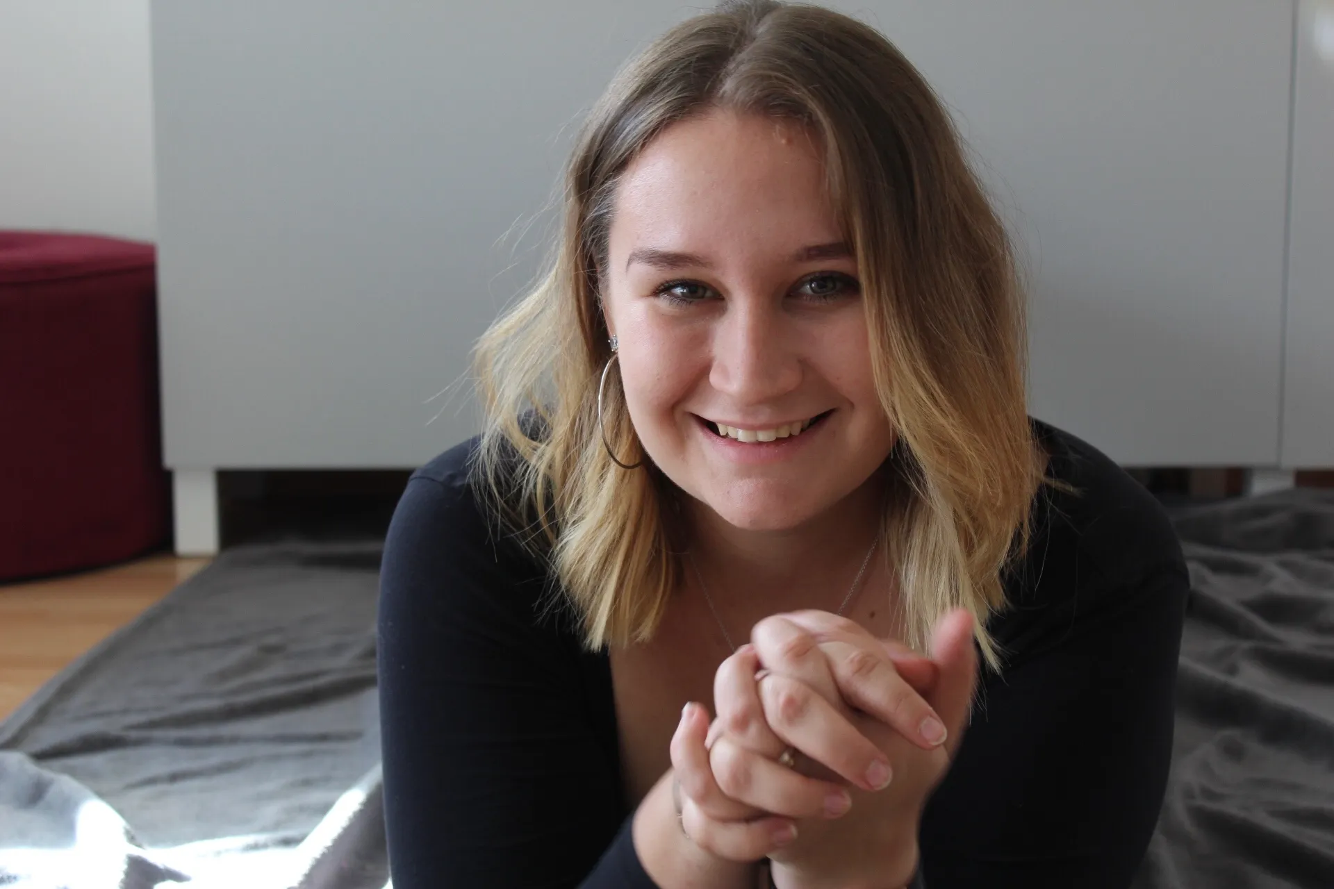 Junge Frau mit schulterlangem blonden Haar lächelt in die Kamera, während sie die Hände vor sich verschränkt, sitzend auf einem grauen Teppich vor einem weißen Möbelstück.