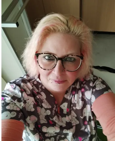 Frau mit kurzen platinblonden Haaren mit rosafarbenen Strähnen, trägt große Brille und ein dunkles Oberteil mit einem weißen Elefantenmuster, vor einer hellen Wand und Holzmöbeln.