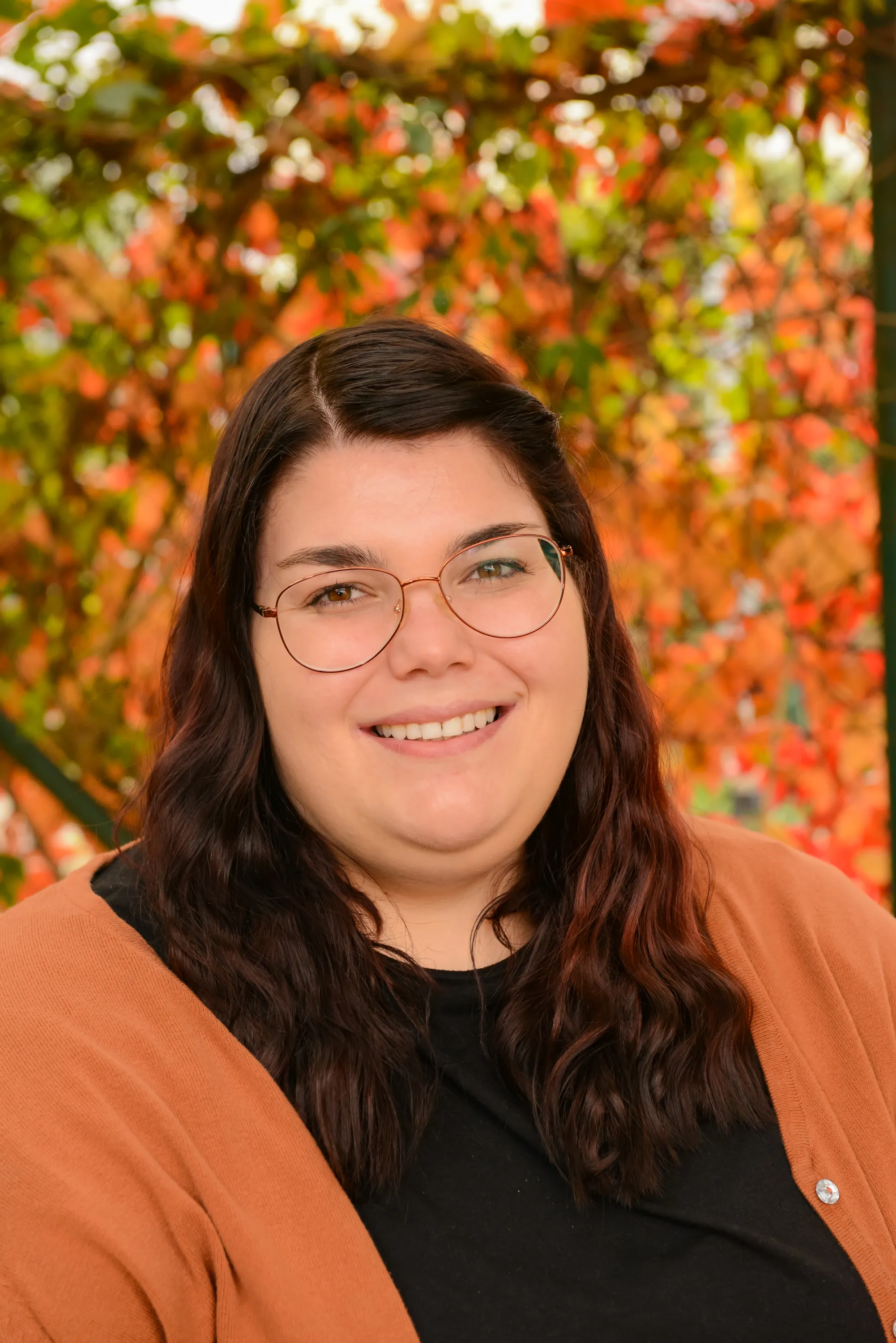 Porträt einer lächelnden Frau mit langen braunen welligen Haaren, die eine Brille trägt und eine schwarze Bluse mit orangem Cardigan vor herbstlichem Hintergrund mit bunten Blättern.