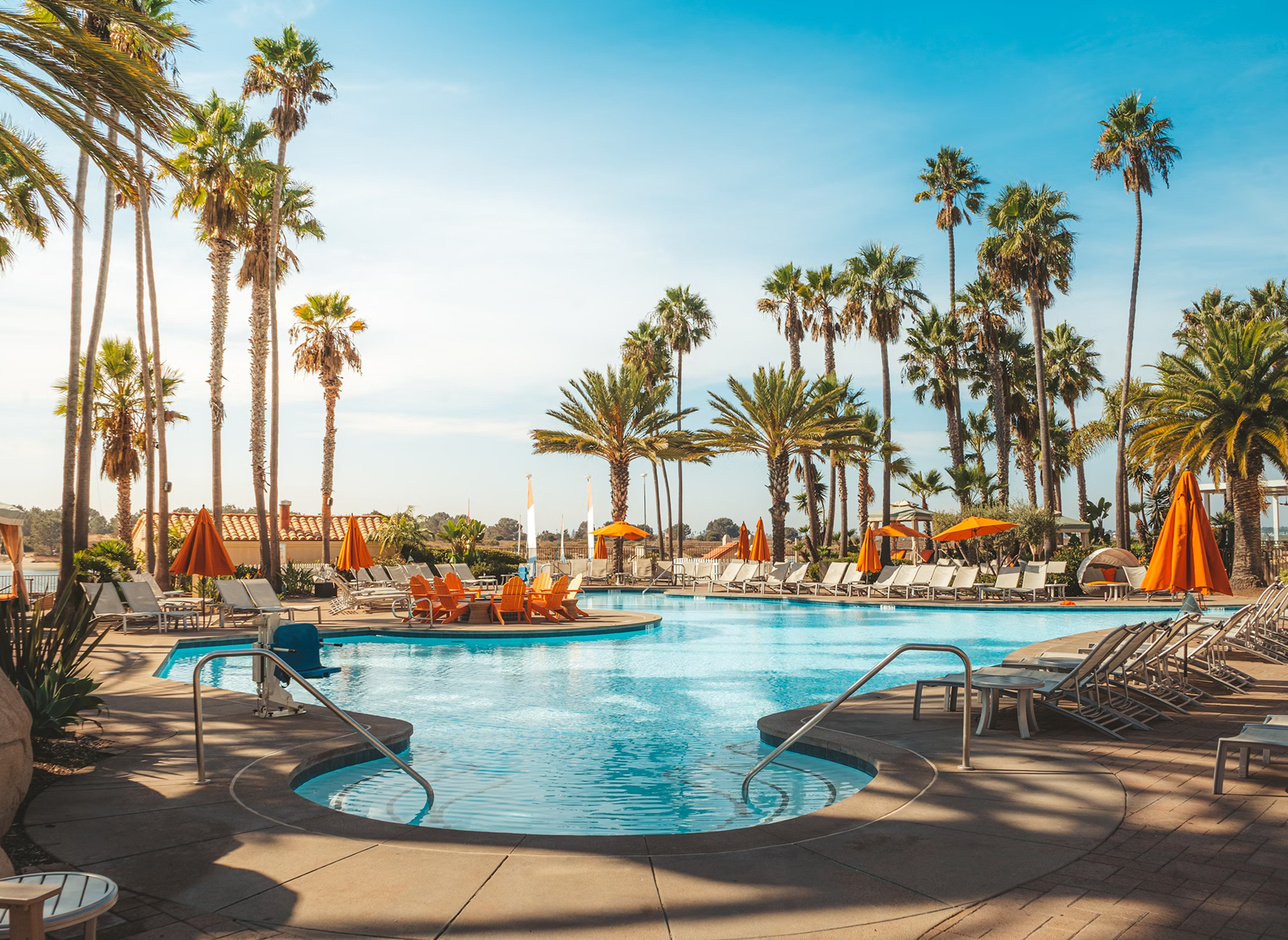 Outdoor swimming pool surrounded by lounge chairs, orange umbrellas, and tall palm trees under a clear blue sky.