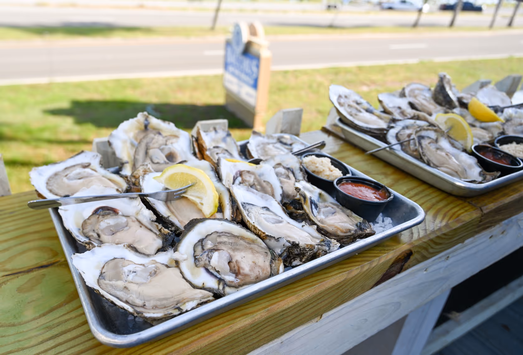 Two trays of freshly shucked oysters served on a wooden counter with lemon wedges and small cups of cocktail sauce and horseradish.