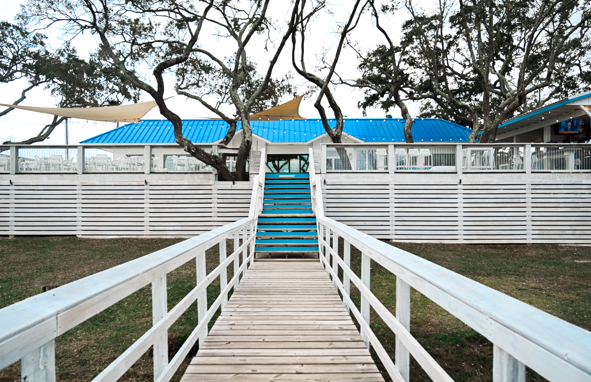 Wooden boardwalk and stairs leading to a white building with a blue roof surrounded by trees and outdoor seating.