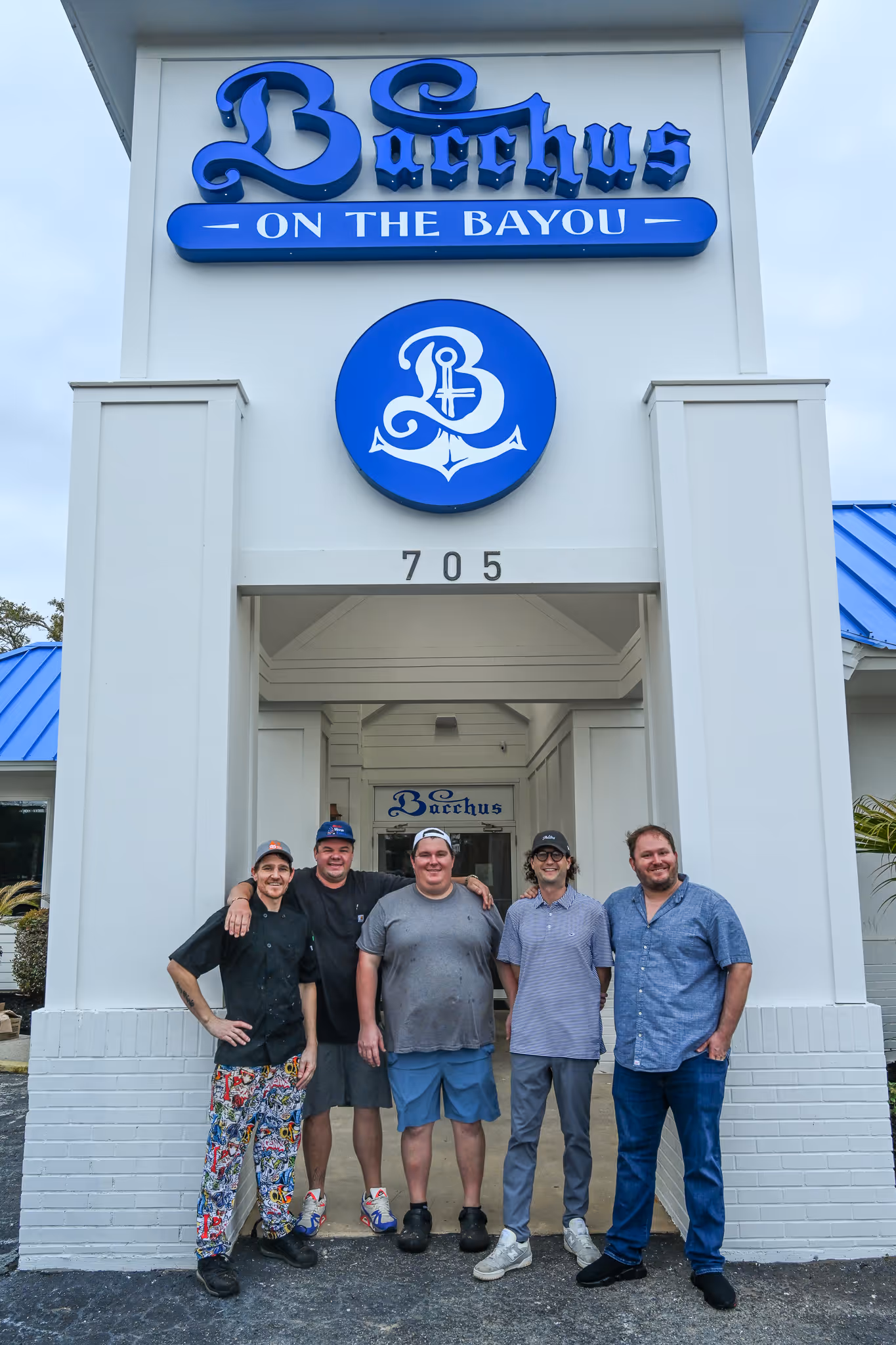 Five men standing and smiling in front of the Bacchus on the Bayou entrance with a large blue sign and the number 705 above them.