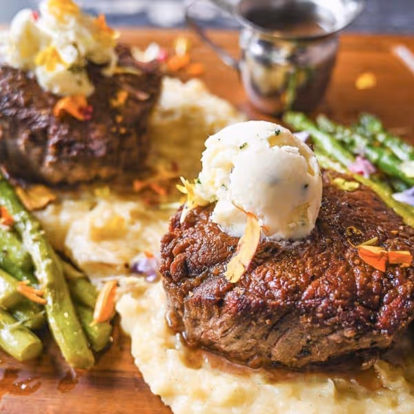 Two cooked filet mignon steaks topped with herb butter, served on creamy mashed potatoes with sautéed asparagus and garnished with edible flowers.