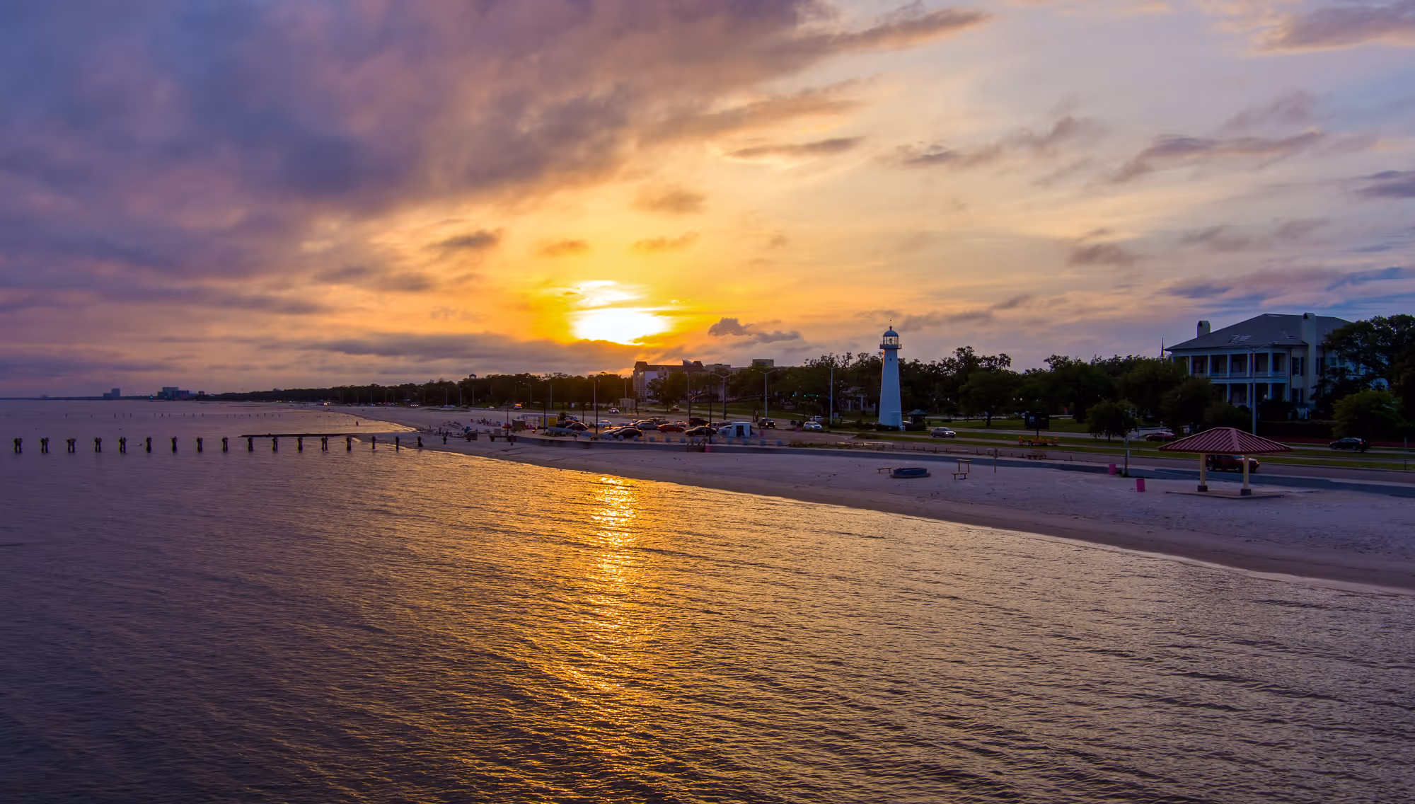 Sunset over a beach with a white lighthouse near a road and buildings in the background.