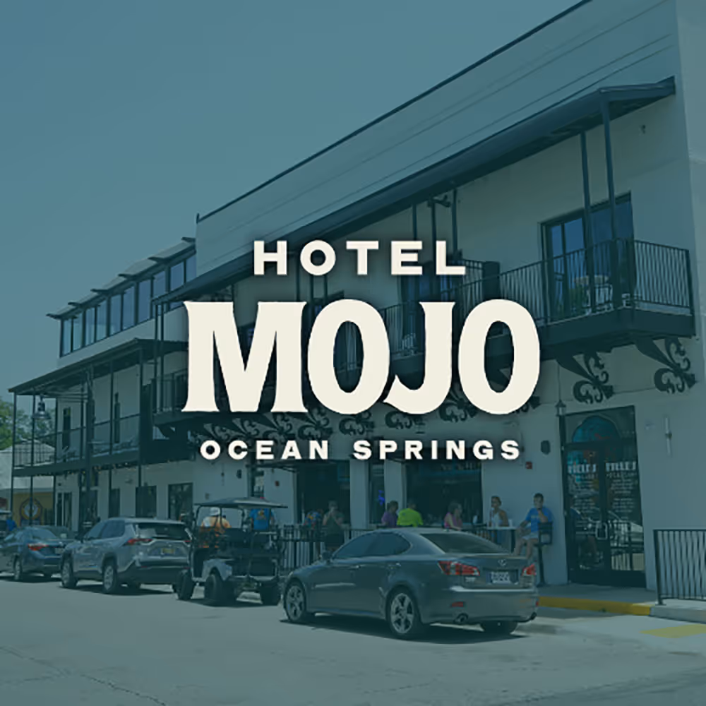 Street view of Hotel Mojo Ocean Springs with parked cars and outdoor seating under balcony railings.