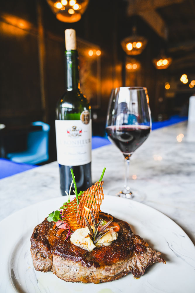 Grilled steak topped with herb butter and garnishes on a white plate, with a bottle of red wine and a glass of wine in the background.