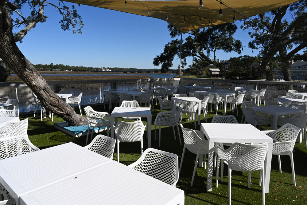 Outdoor dining area with white tables and chairs on green turf overlooking a calm bayou under a clear blue sky.